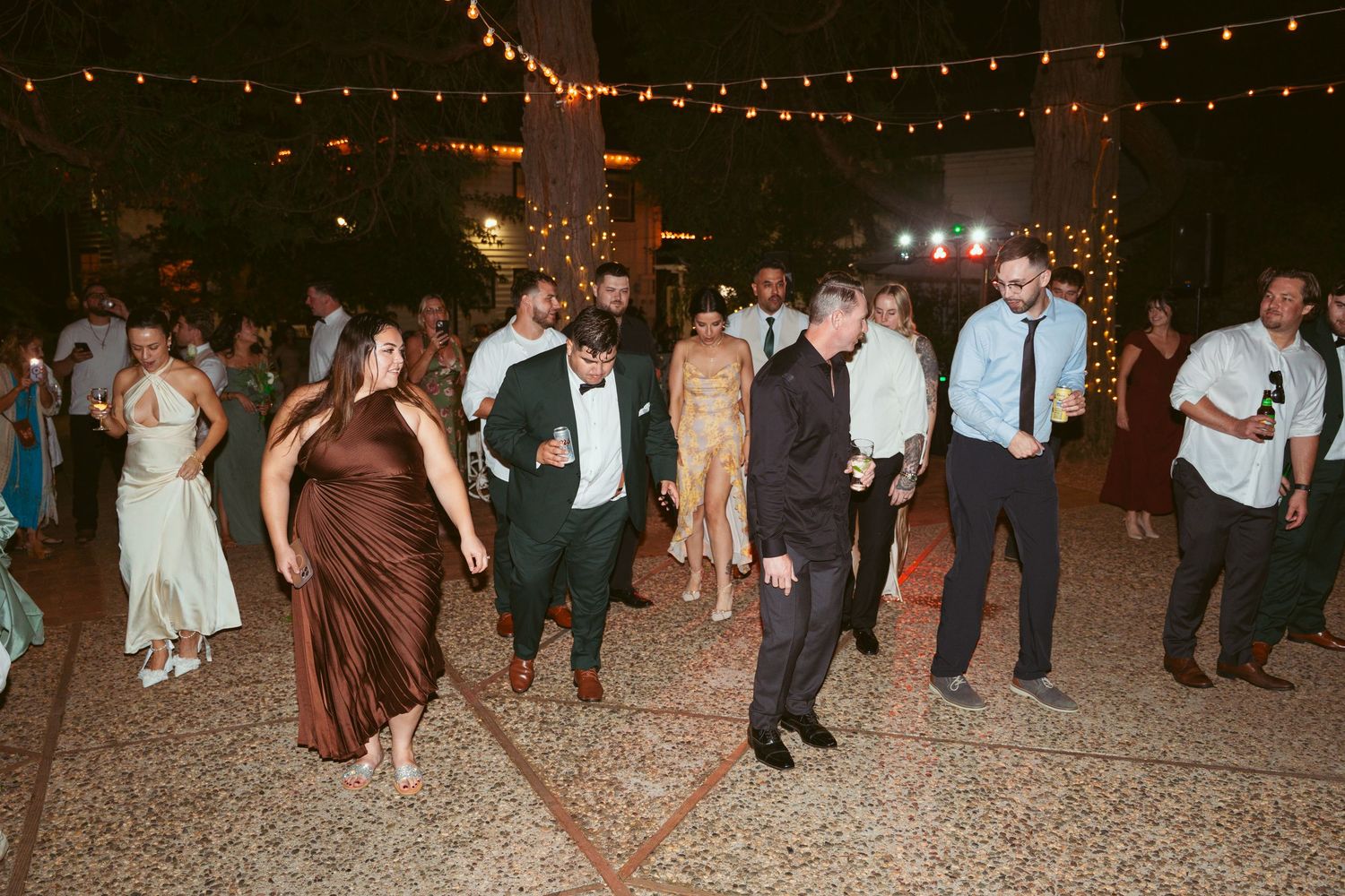 Large group of wedding guests dancing on an outdoor patio under string lights.