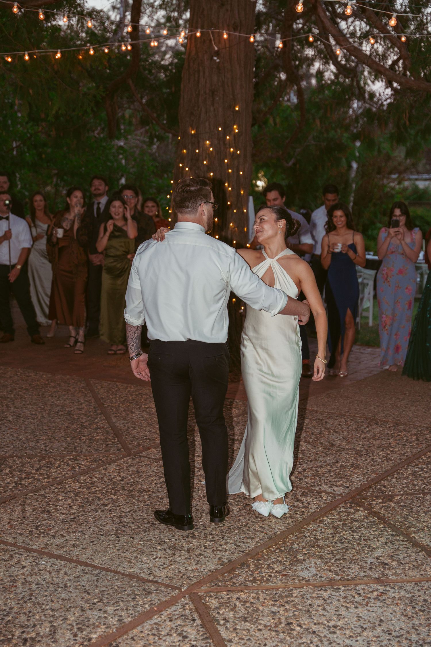 Couples dance together at an outdoor evening reception under string lights and tall trees.