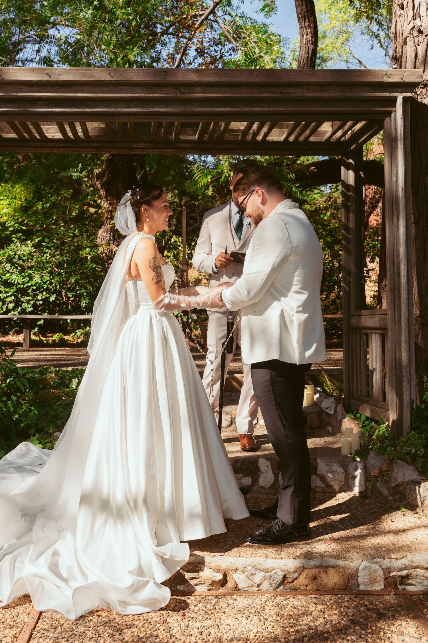 Bride in flowing white gown and groom in white tuxedo share ceremony moment under rustic pergola.