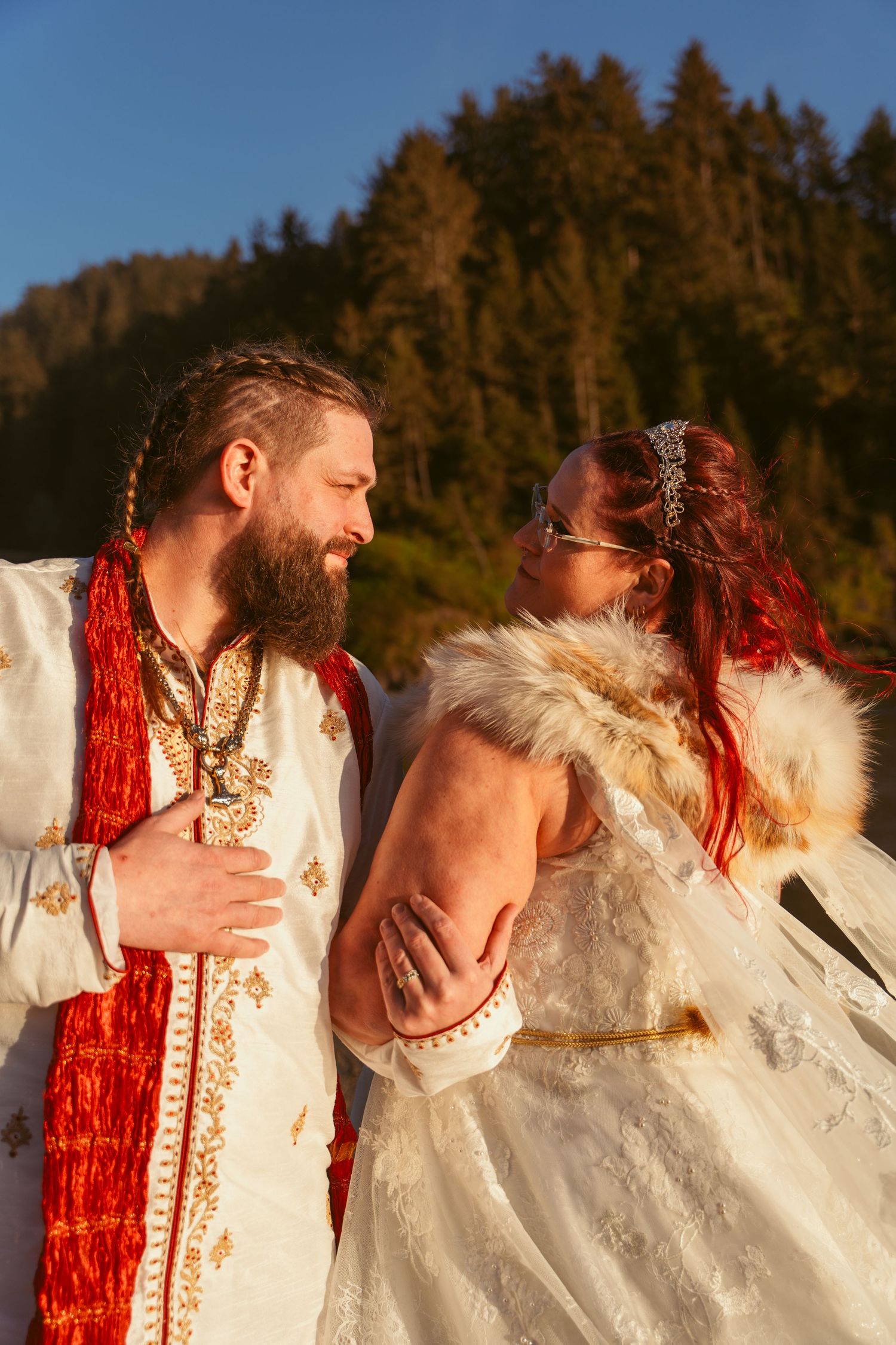 Wedding couple in white and red attire embracing outdoors with evergreen trees in background.