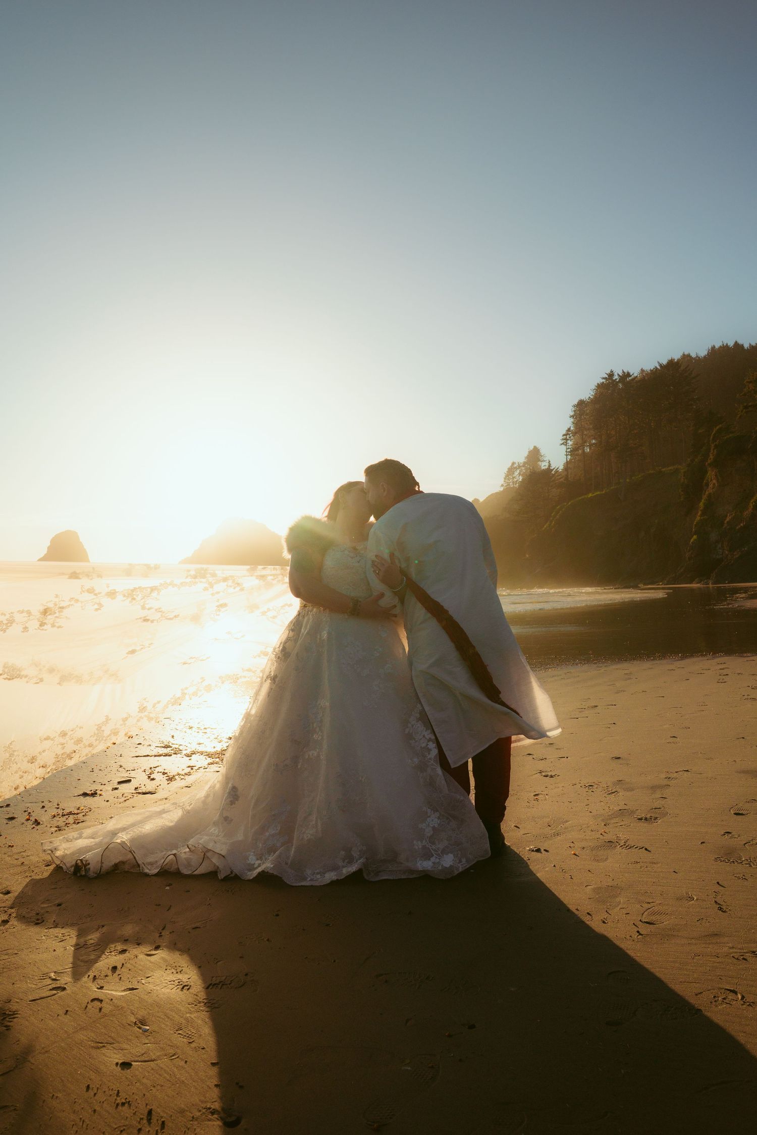 A couple in wedding attire shares an intimate moment on a beach during golden hour.