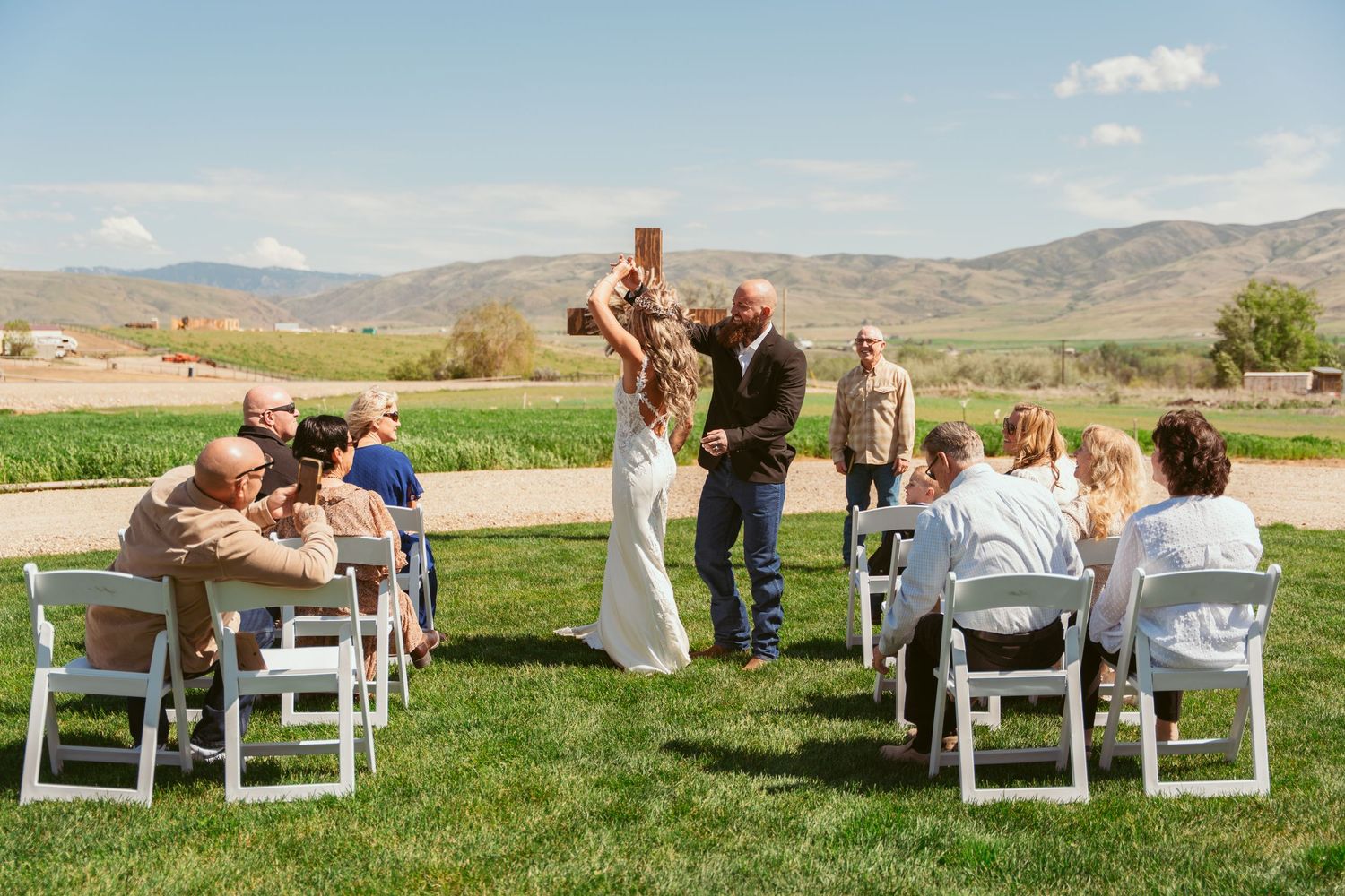 An intimate wedding ceremony unfolds on a grassy lawn with guests seated in white chairs.
