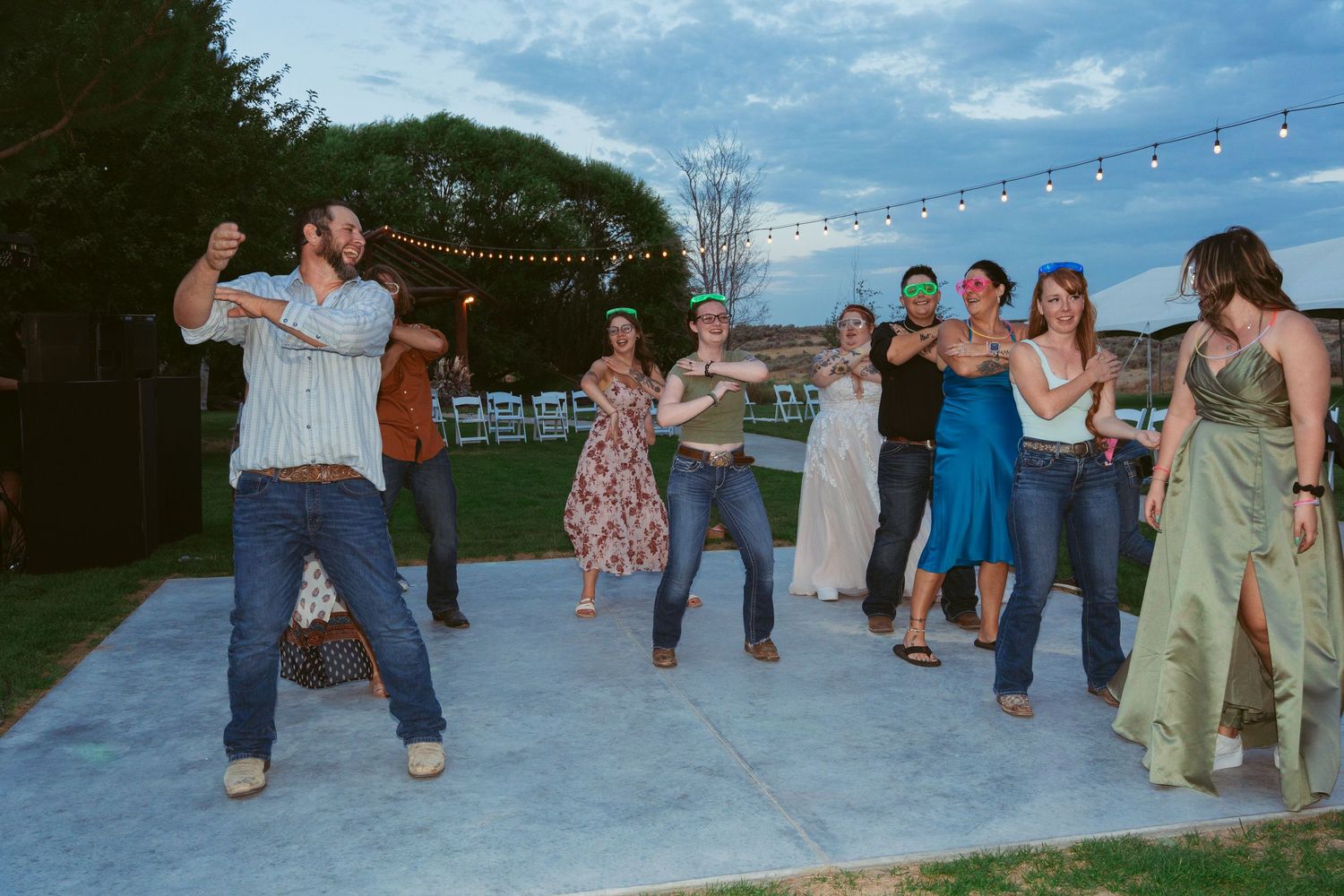 Wedding guests dance and socialize outdoors under string lights during sunset.