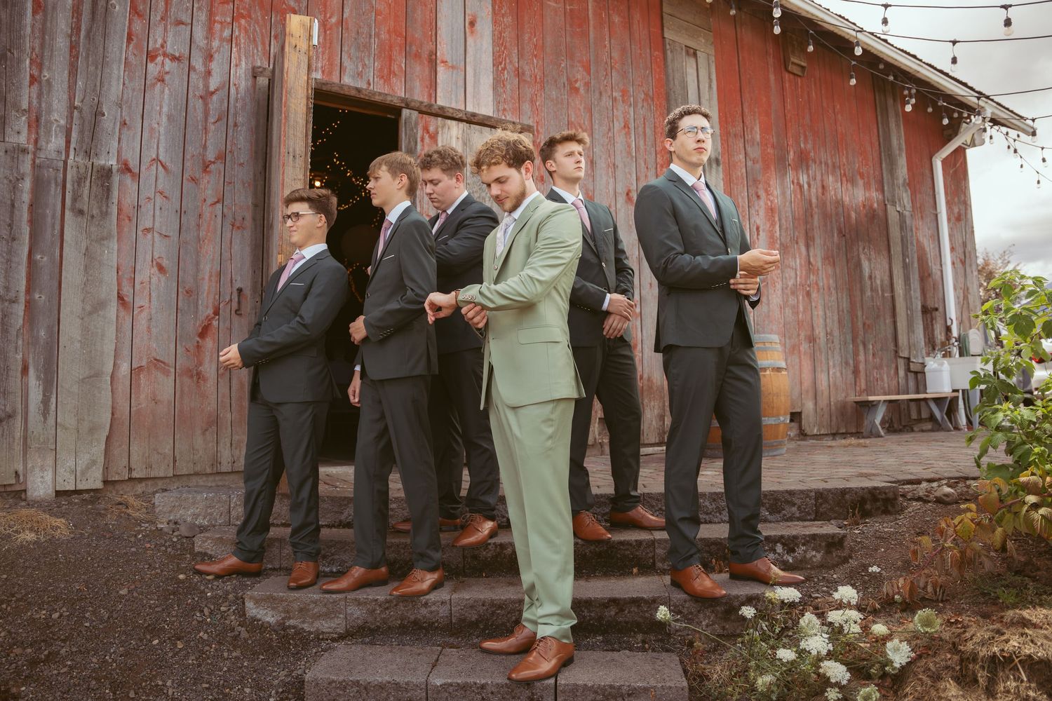 Groomsmen in suits pose together against rustic red barn wall with string lights overhead.
