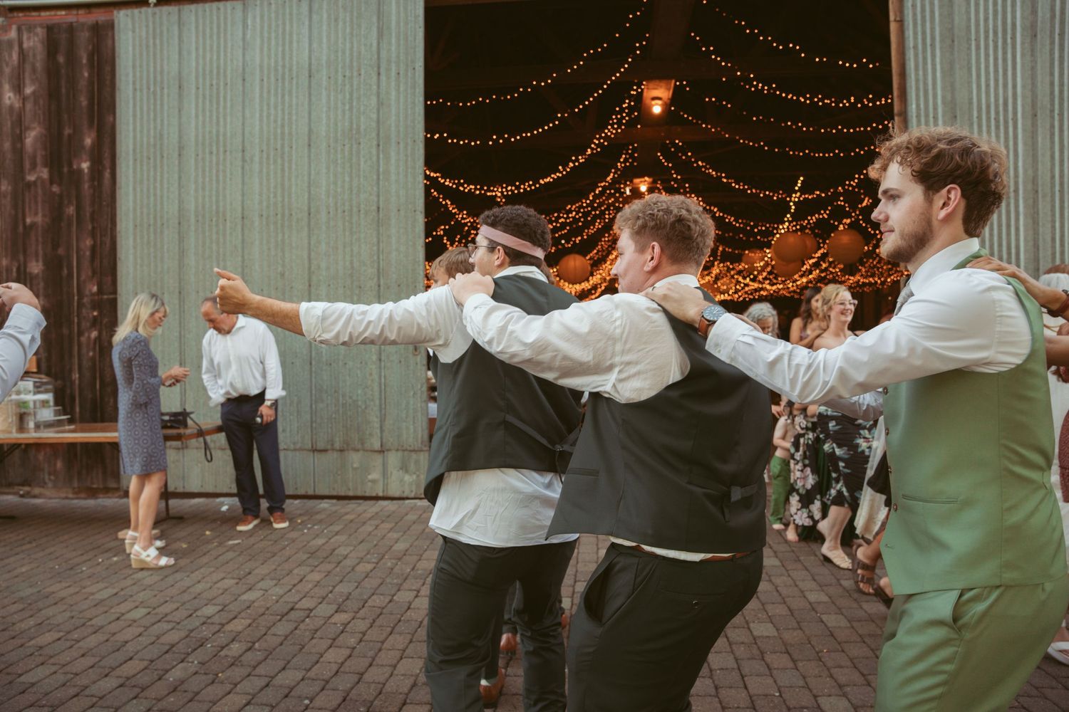 Group dance routine entertains guests at rustic outdoor wedding venue with string lights.