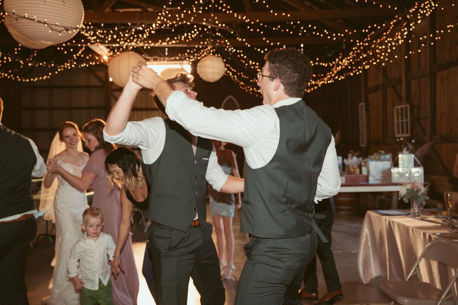 Guests dance and celebrate under glowing string lights and paper lanterns at a rustic wedding reception.