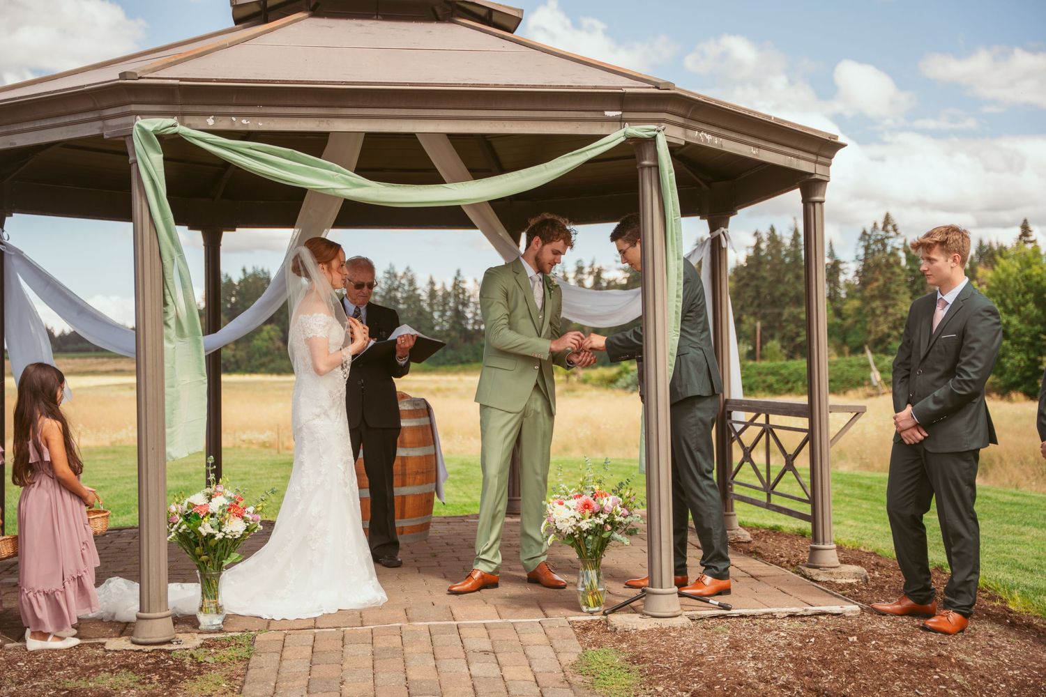 Outdoor wedding ceremony under rustic gazebo with pink flowers and white draping.