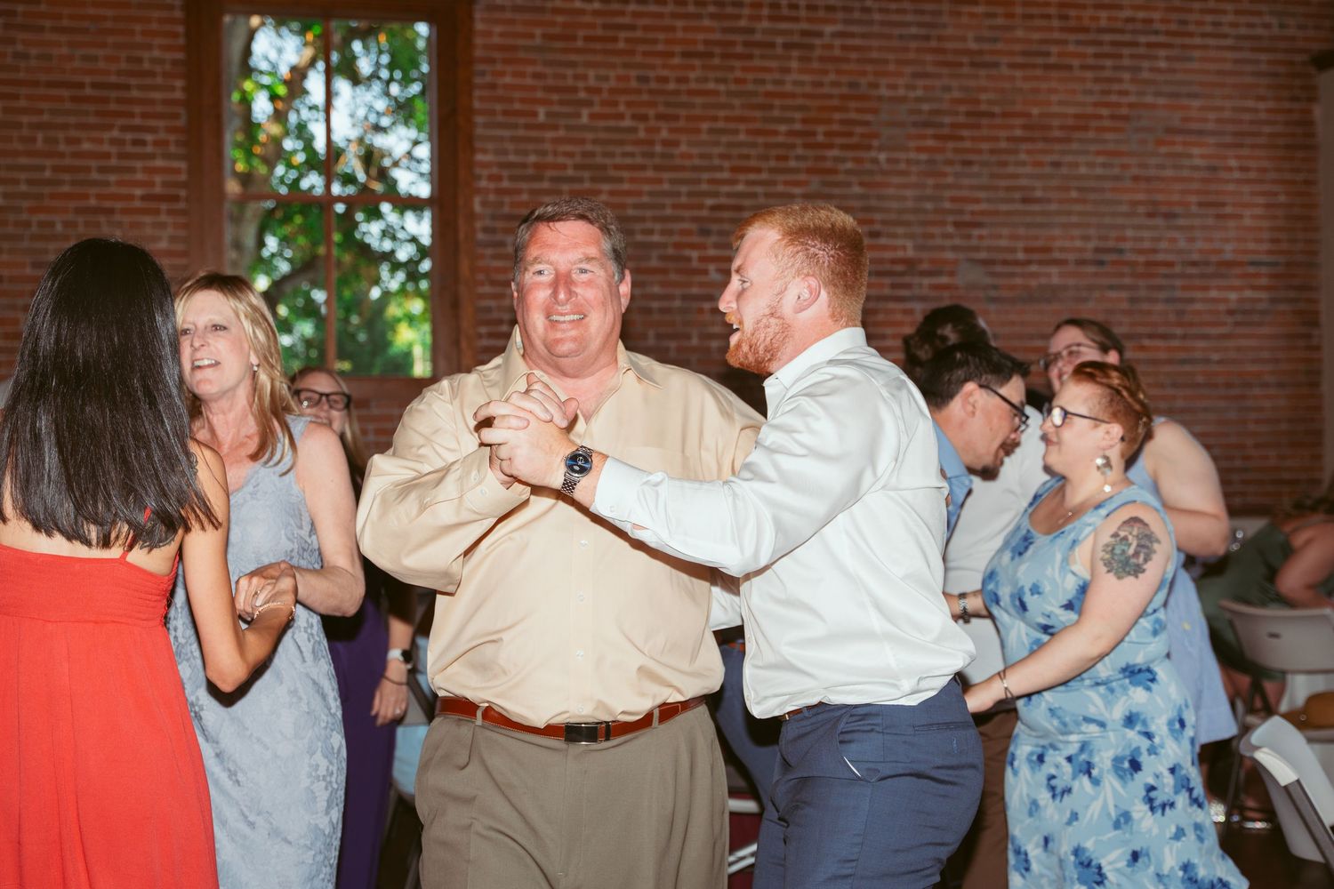Wedding guests dance and celebrate together in a brick-walled reception venue with string lights.
