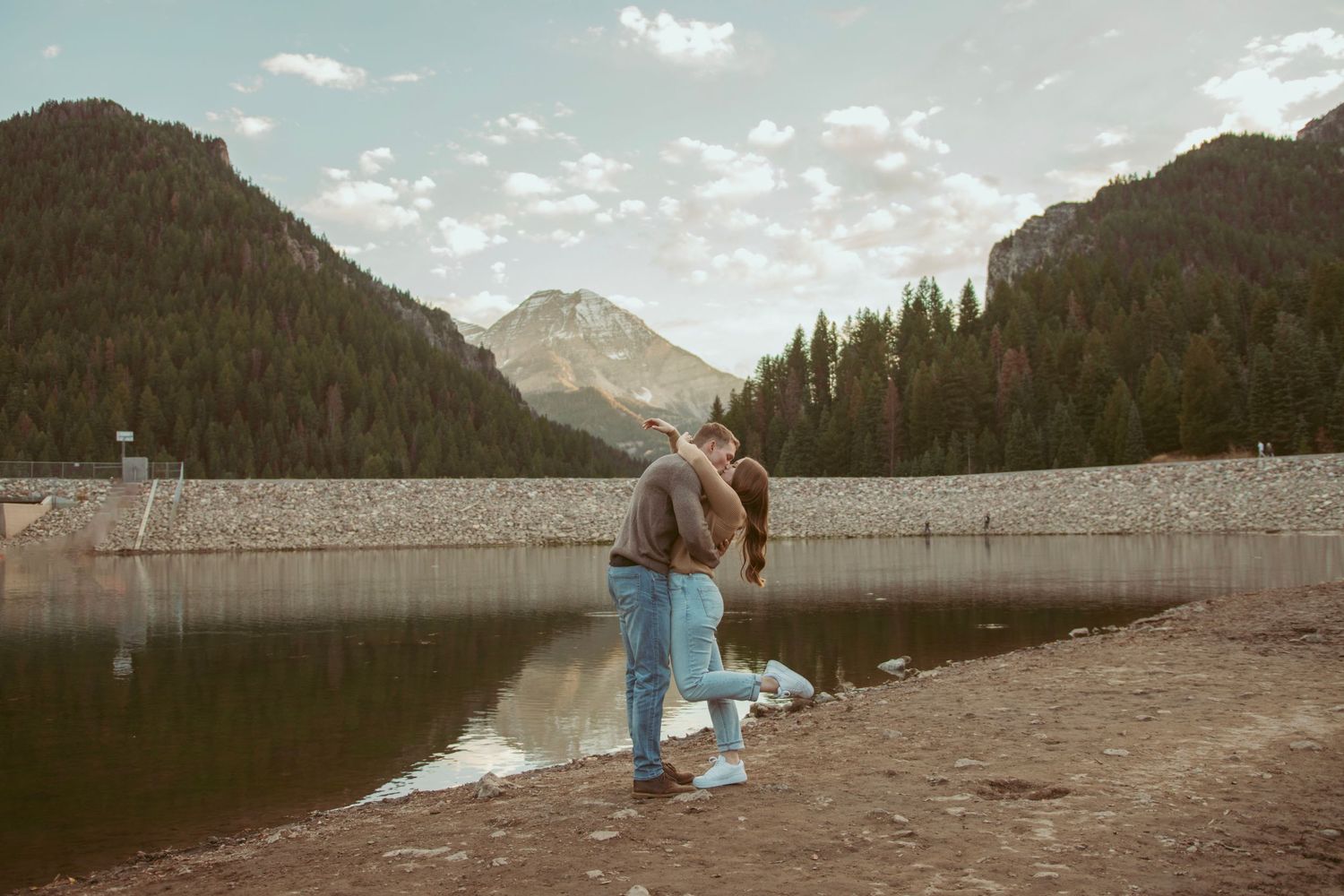 Couple embracing on the shore of a mountain reservoir with evergreen forests in the distance.
