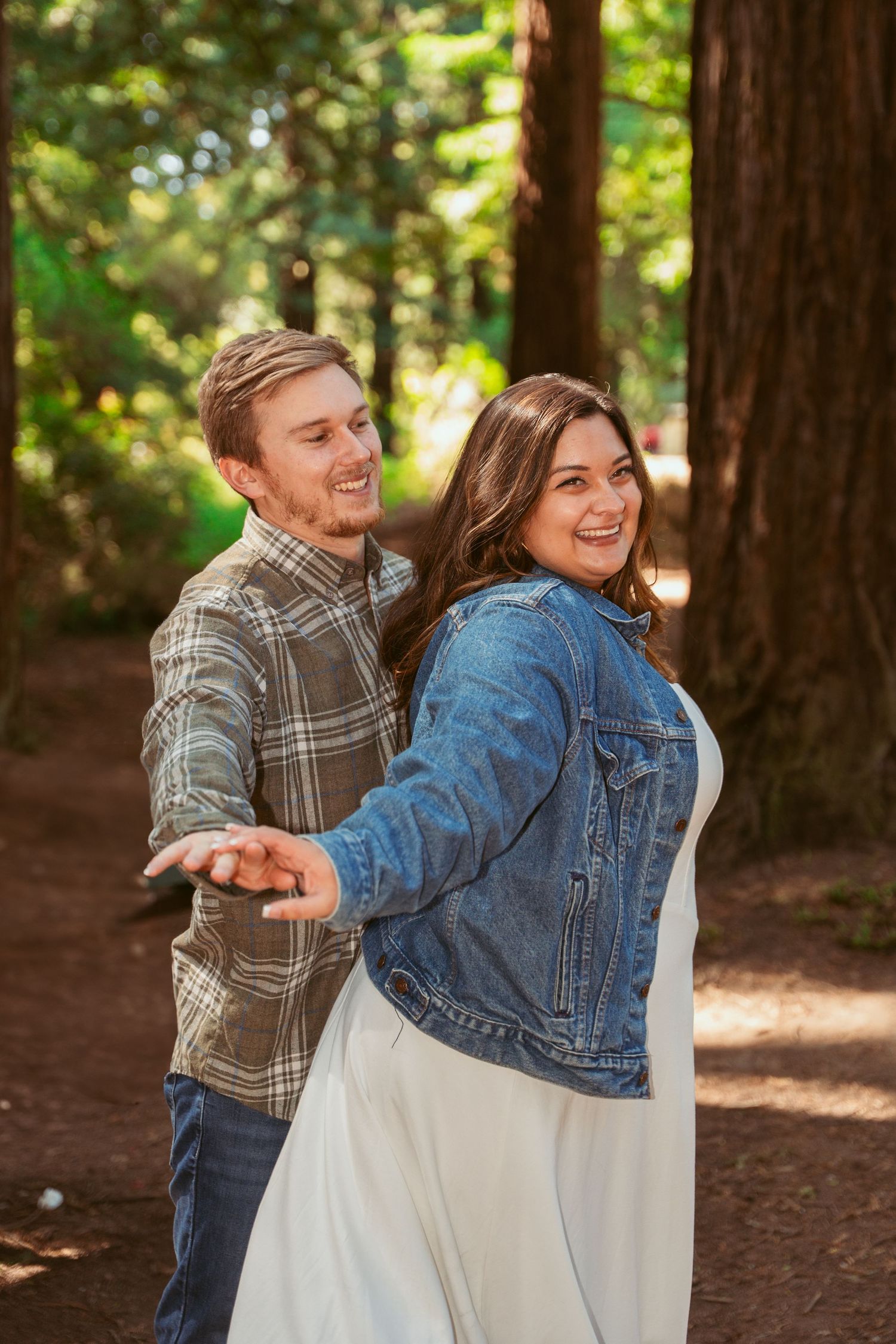 A couple laughs together while sharing a playful moment among tall redwood trees.