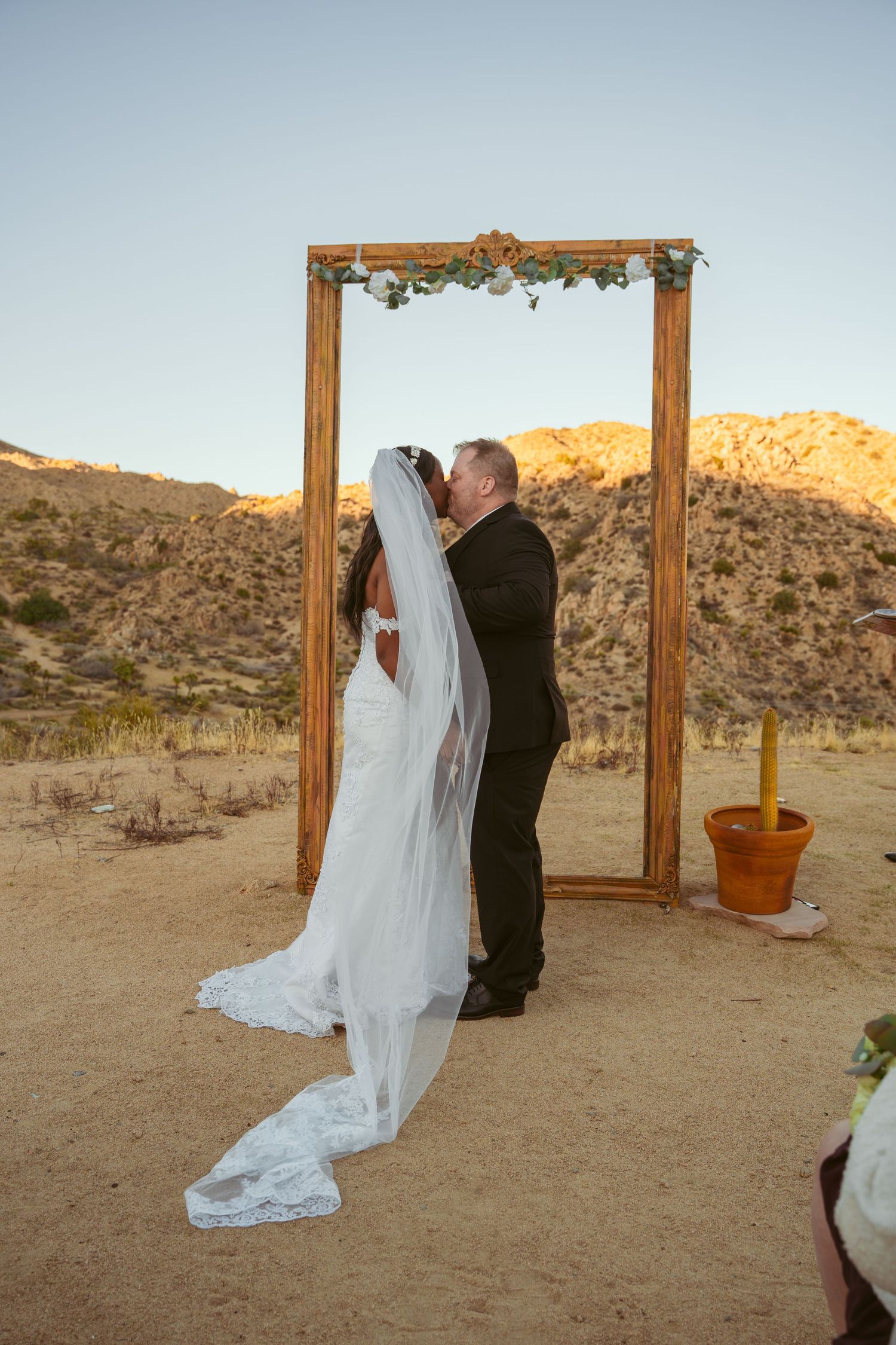 Wedding ceremony in progress under a wooden arch with desert mountains and golden sunlight.