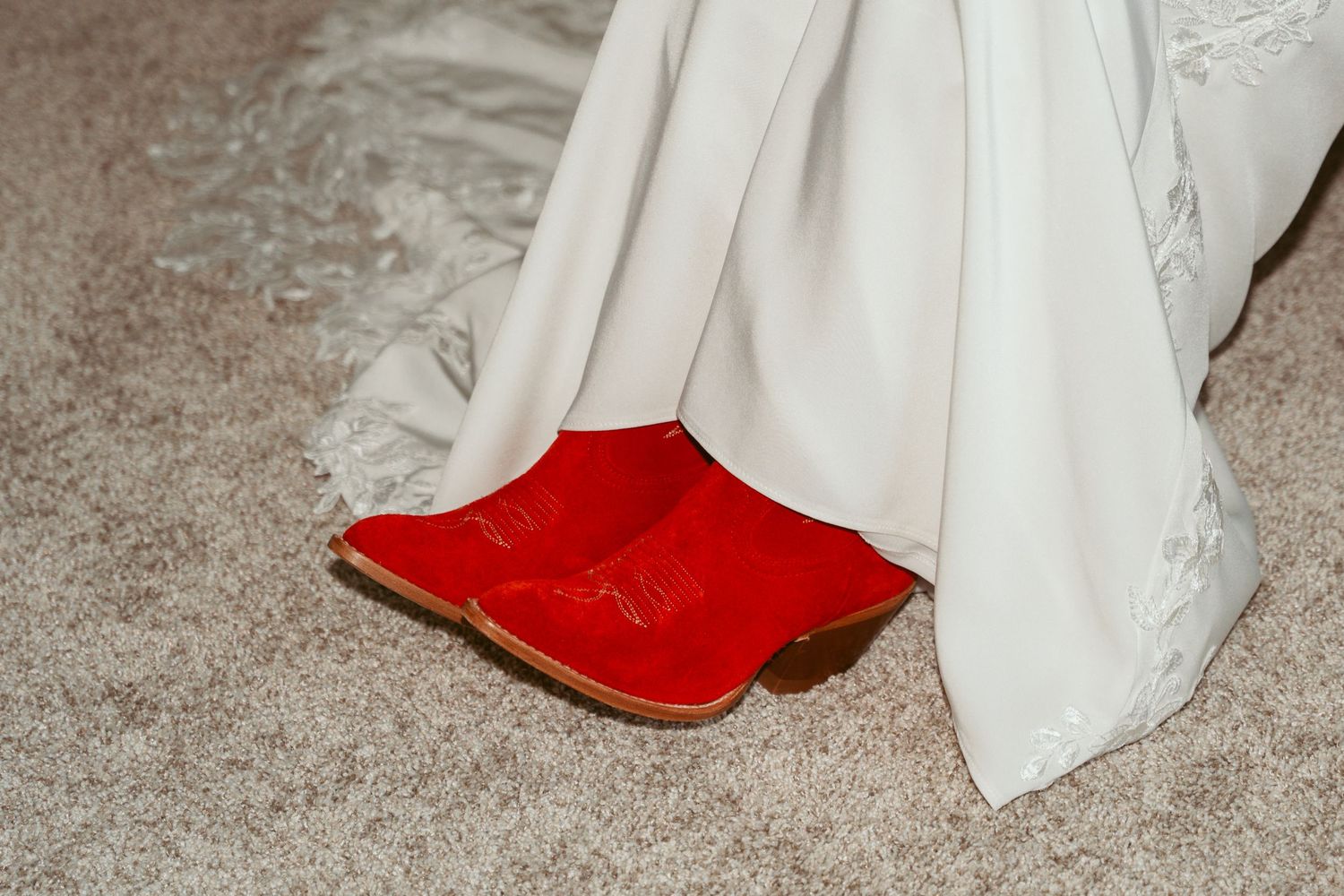 Bright red cowboy boots peeking out from under a white wedding dress on gray carpet.