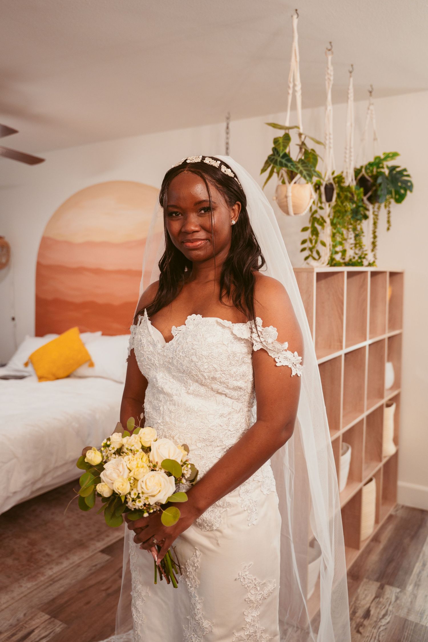 A bride in a white lace wedding gown holds a bouquet of cream roses in an elegantly decorated room.