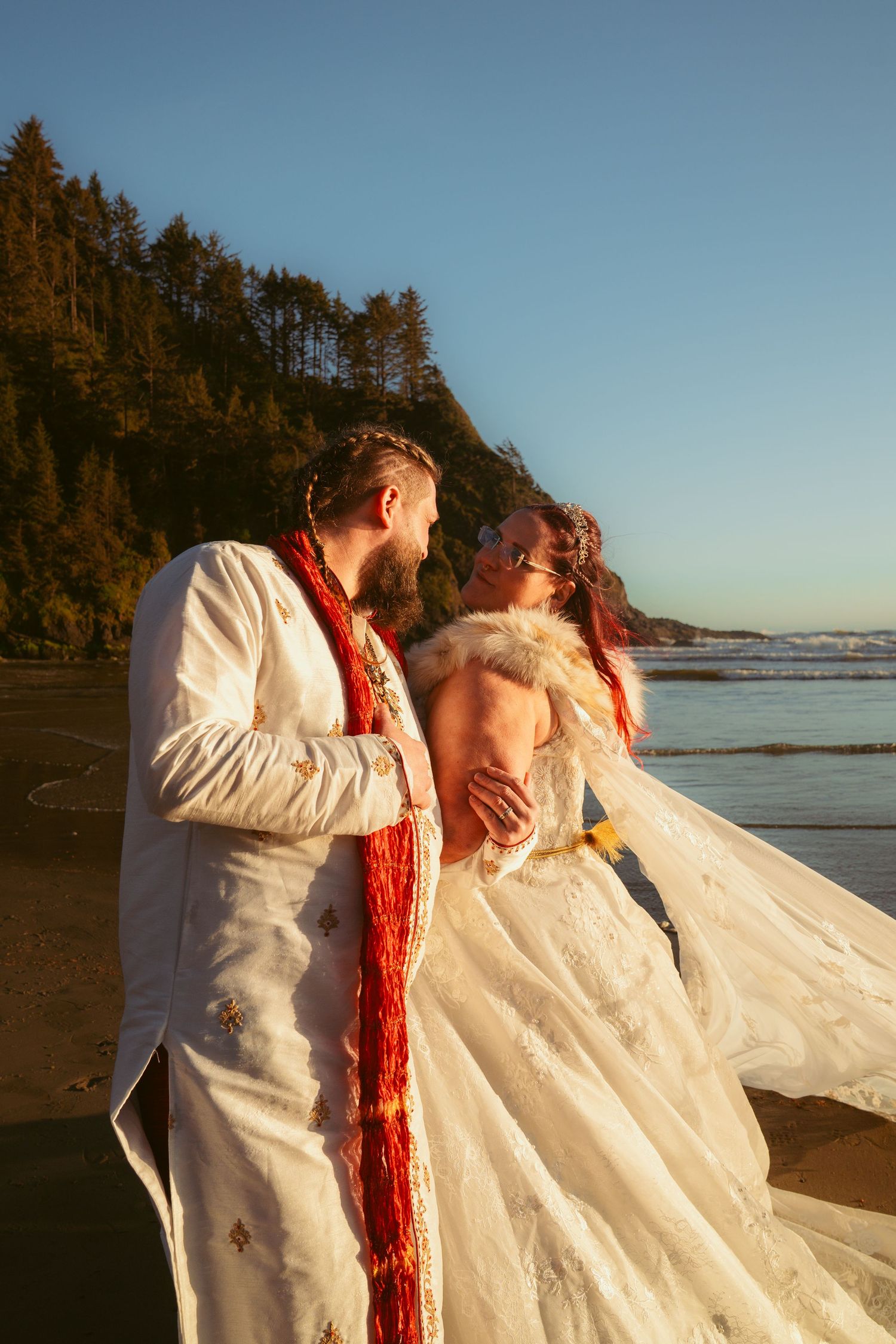 A couple in white wedding attire share a romantic moment on a beach with evergreen trees in the background.