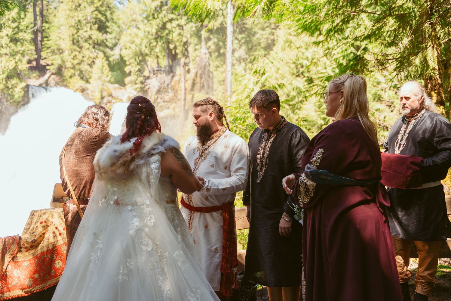 Wedding party and guests gather in a wooded outdoor setting for the ceremony.