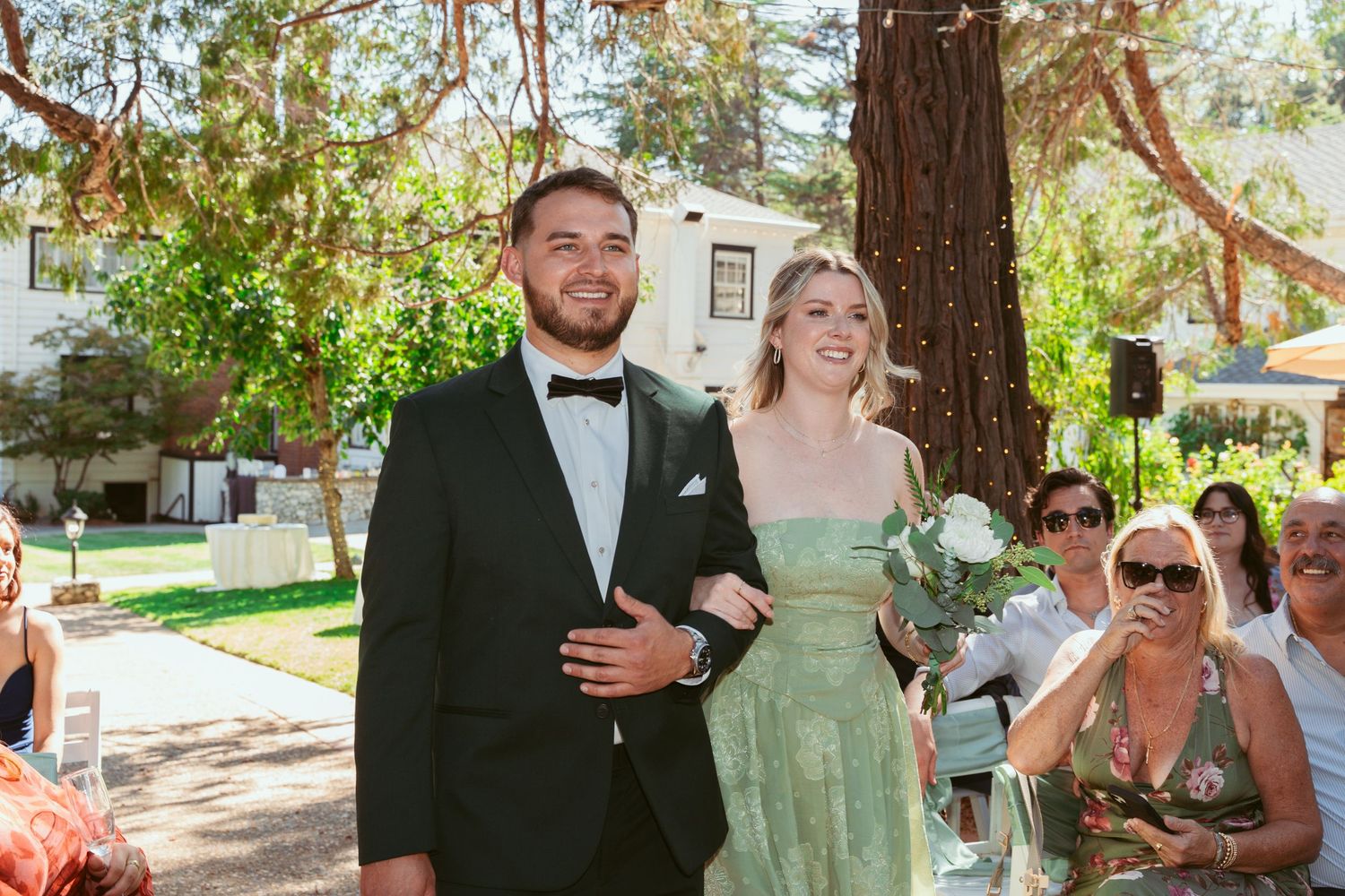 Wedding couple walks down outdoor path while guests look on in sunlit garden setting.
