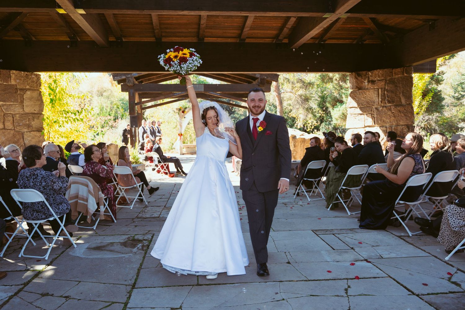 Wedding ceremony setup under a rustic pavilion with white chairs and autumn foliage in background.