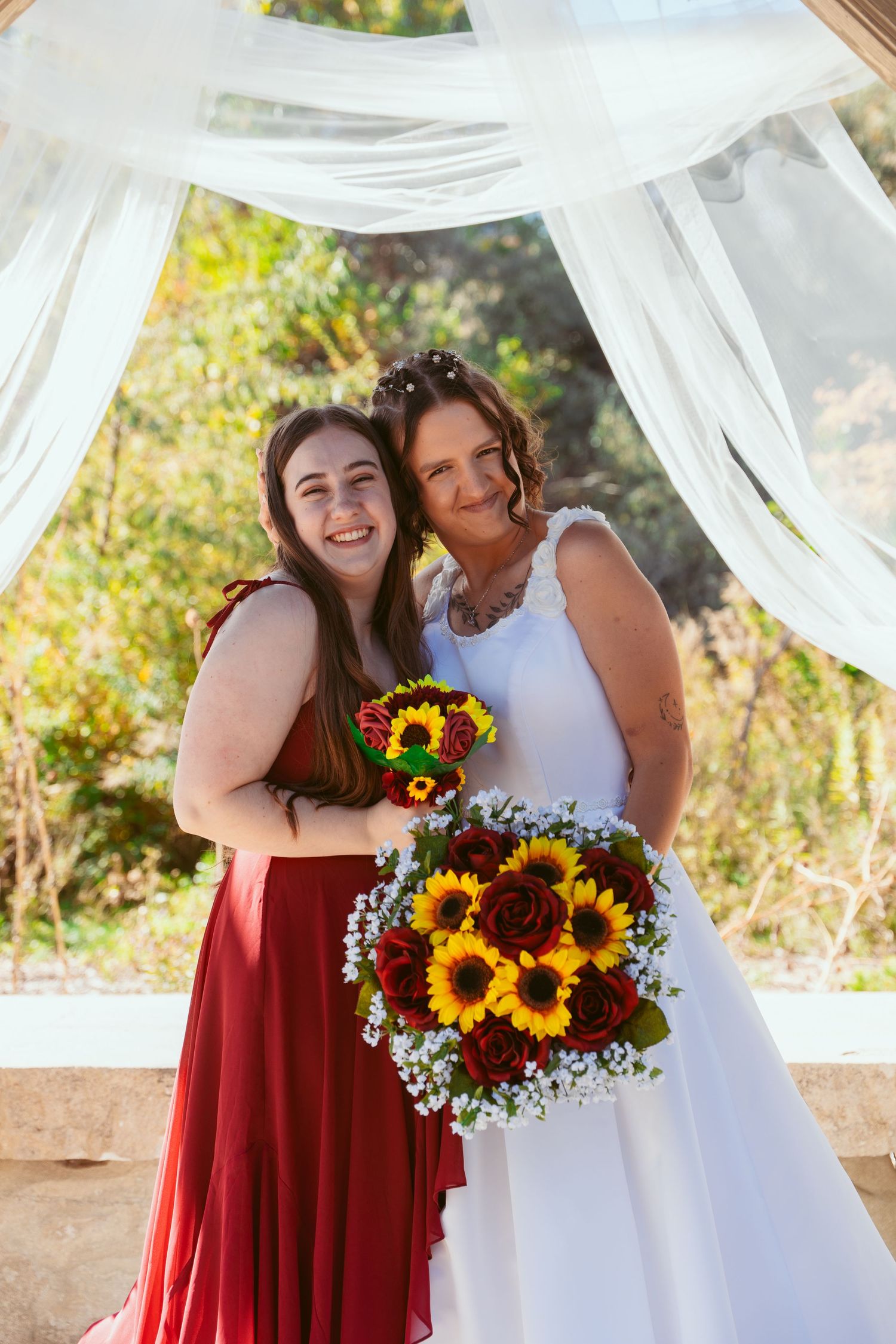 Two people in red and white dresses share a joyful moment with sunflower bouquets under white draping.