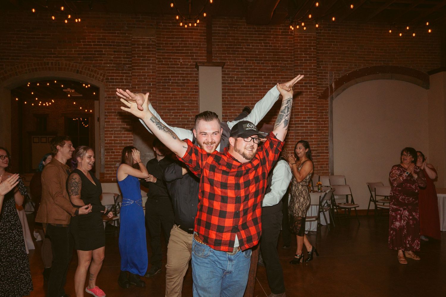 Partygoers dancing enthusiastically under string lights in a rustic brick venue.