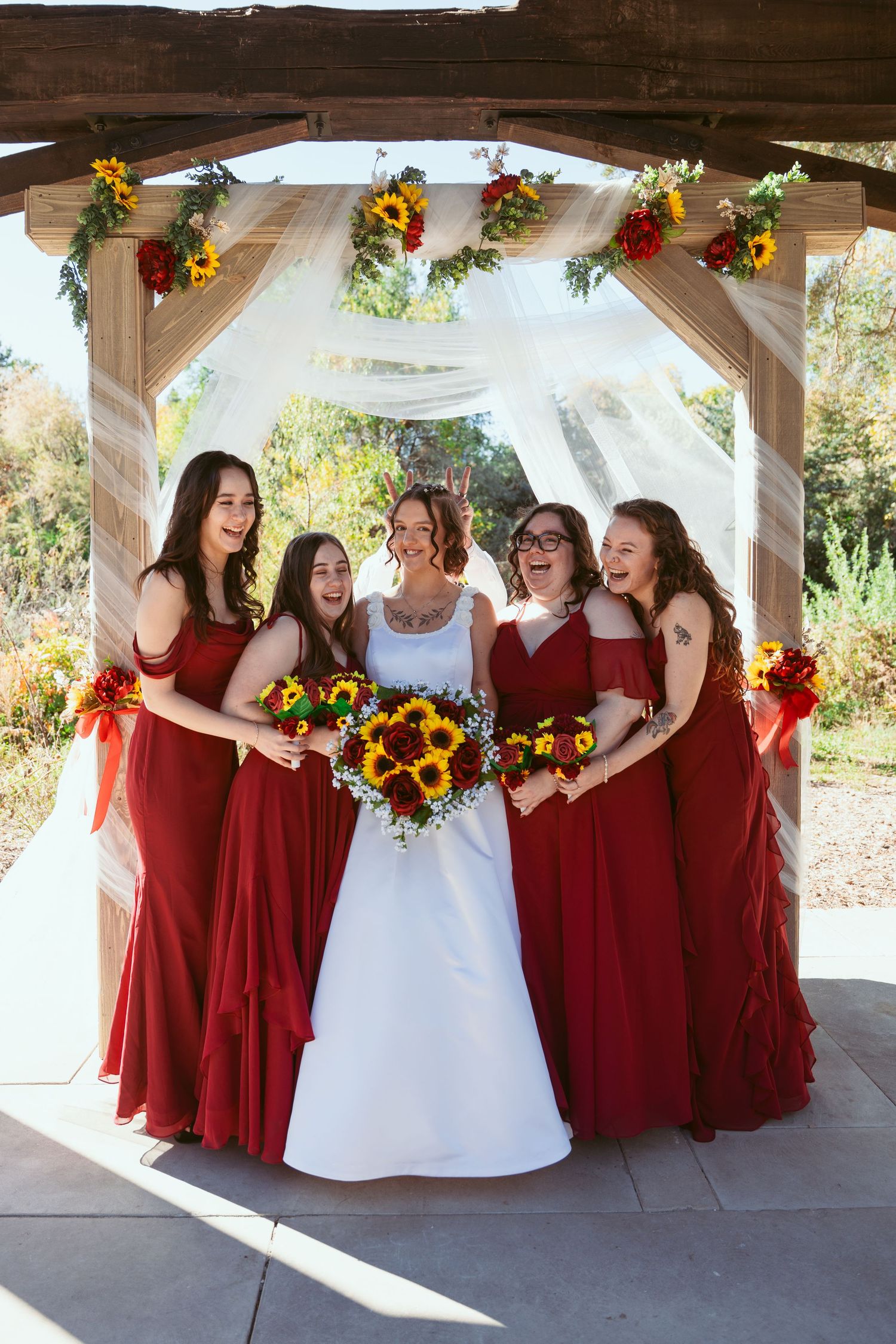 Bridal party in deep red dresses poses with sunflower bouquets against rustic backdrop.
