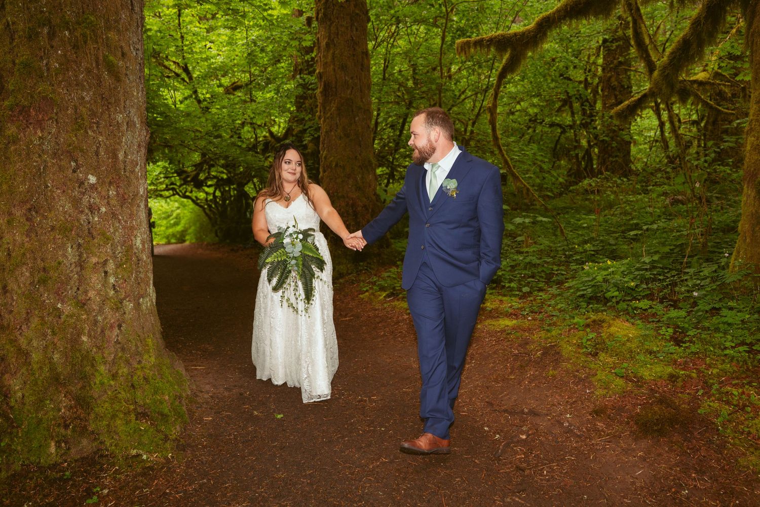 A wedding couple walks hand in hand down a scenic forest trail surrounded by greenery.