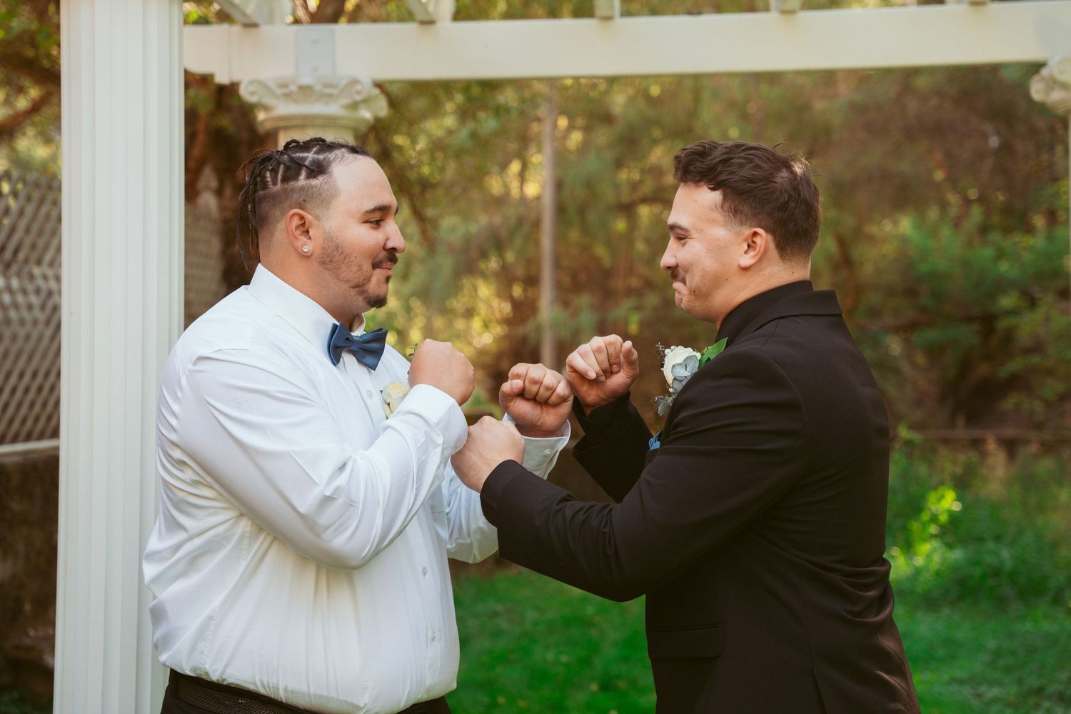 Two people in formal attire playfully interact under a white pergola in a garden setting.