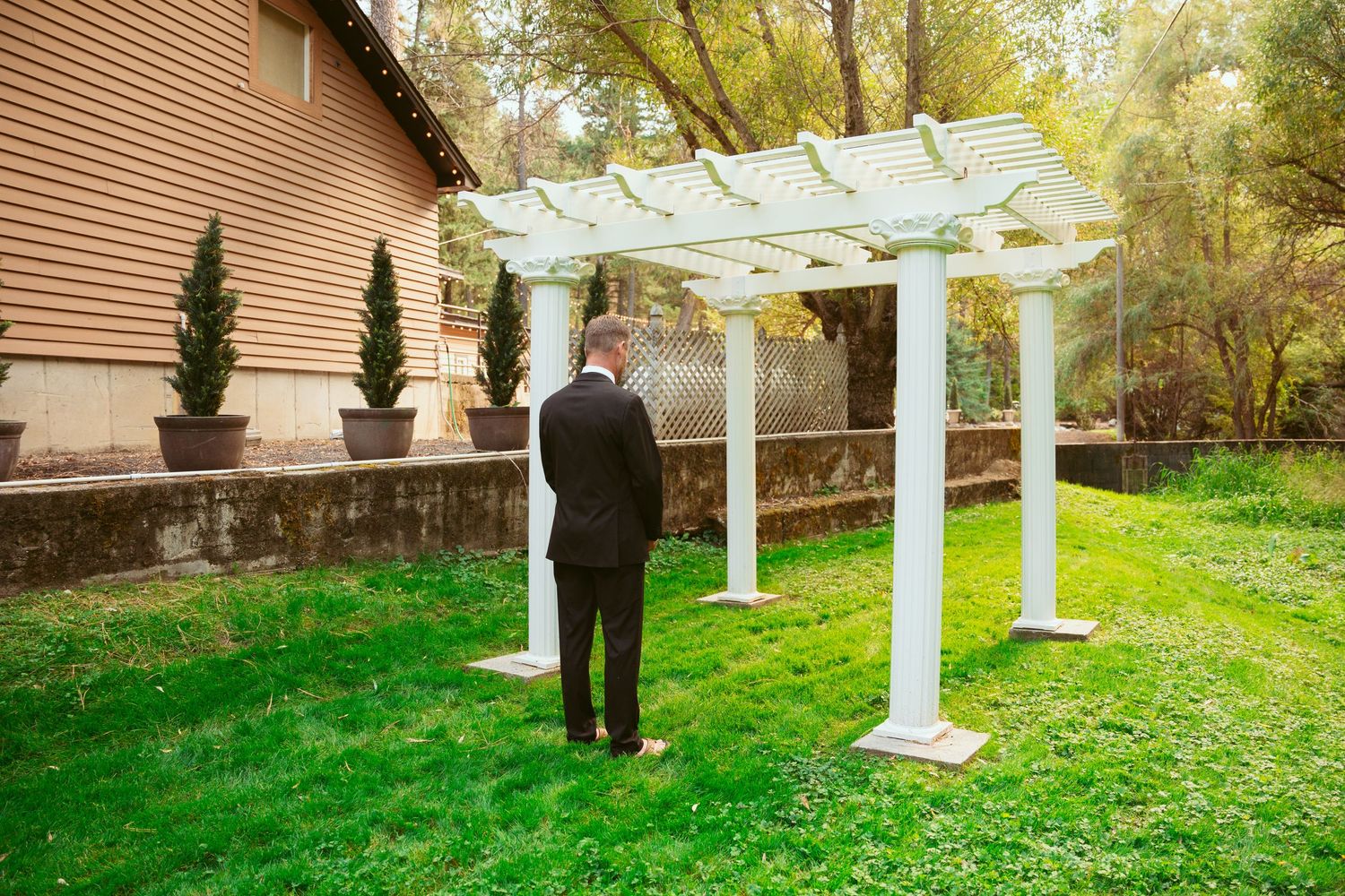 A formal black-suited figure stands alone beneath a white garden pergola on a grassy lawn.