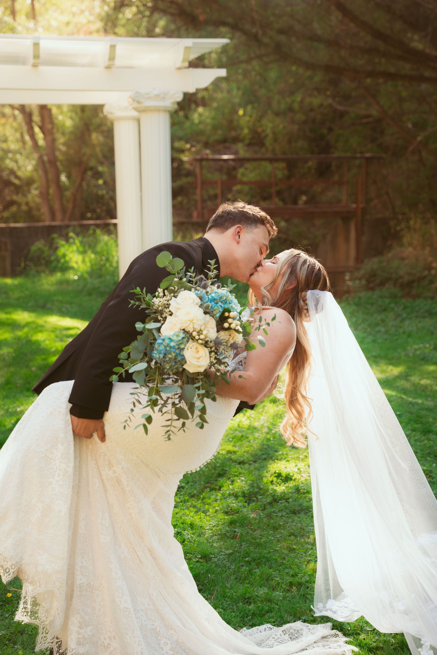 Romantic wedding portrait under white garden pergola with flowing veil and floral bouquet.