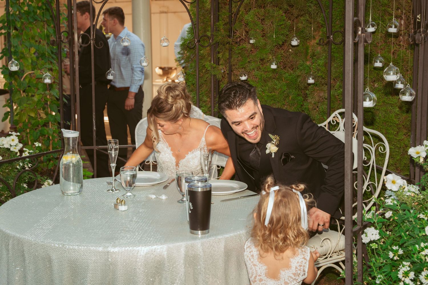 Wedding guests share a moment at an intimate outdoor table setting with twinkling lights and greenery.