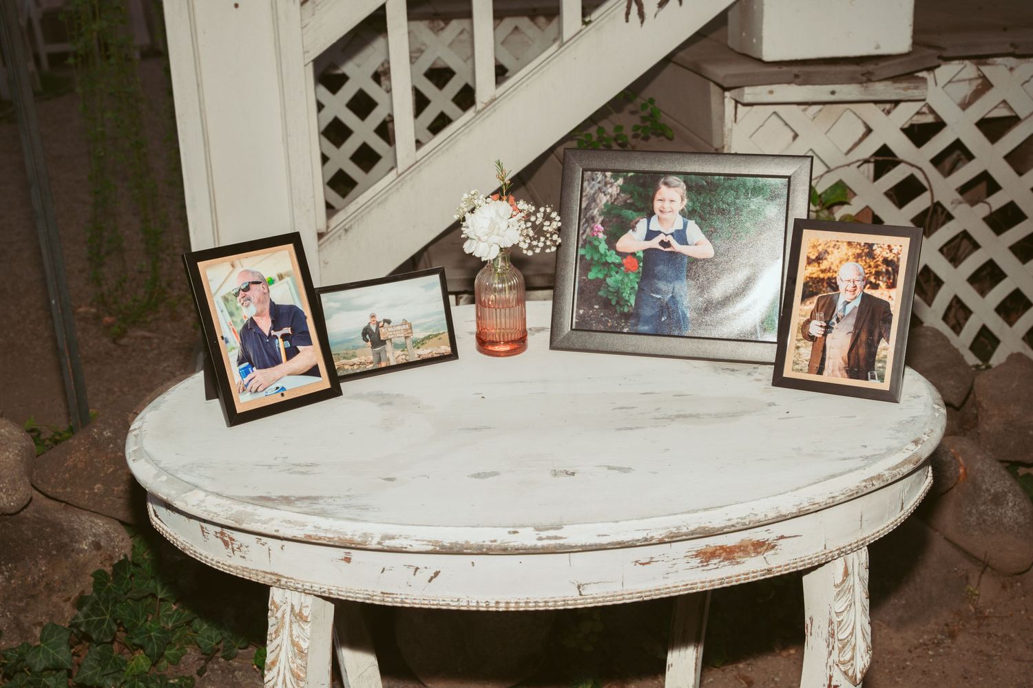 A vintage white round table displays framed photographs and decorative items in a rustic outdoor setting.
