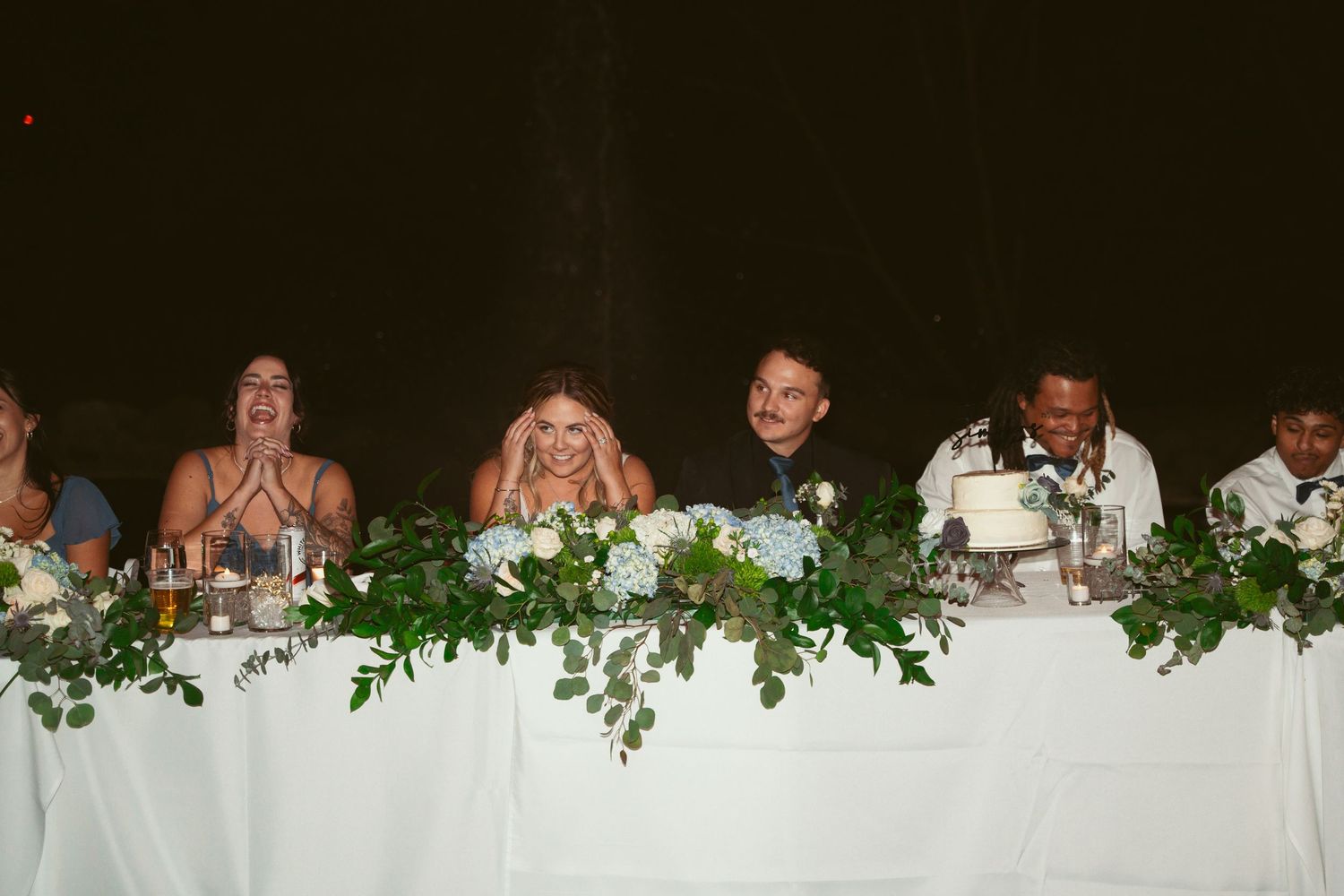 Wedding reception head table decorated with greenery and white linens under string lights.
