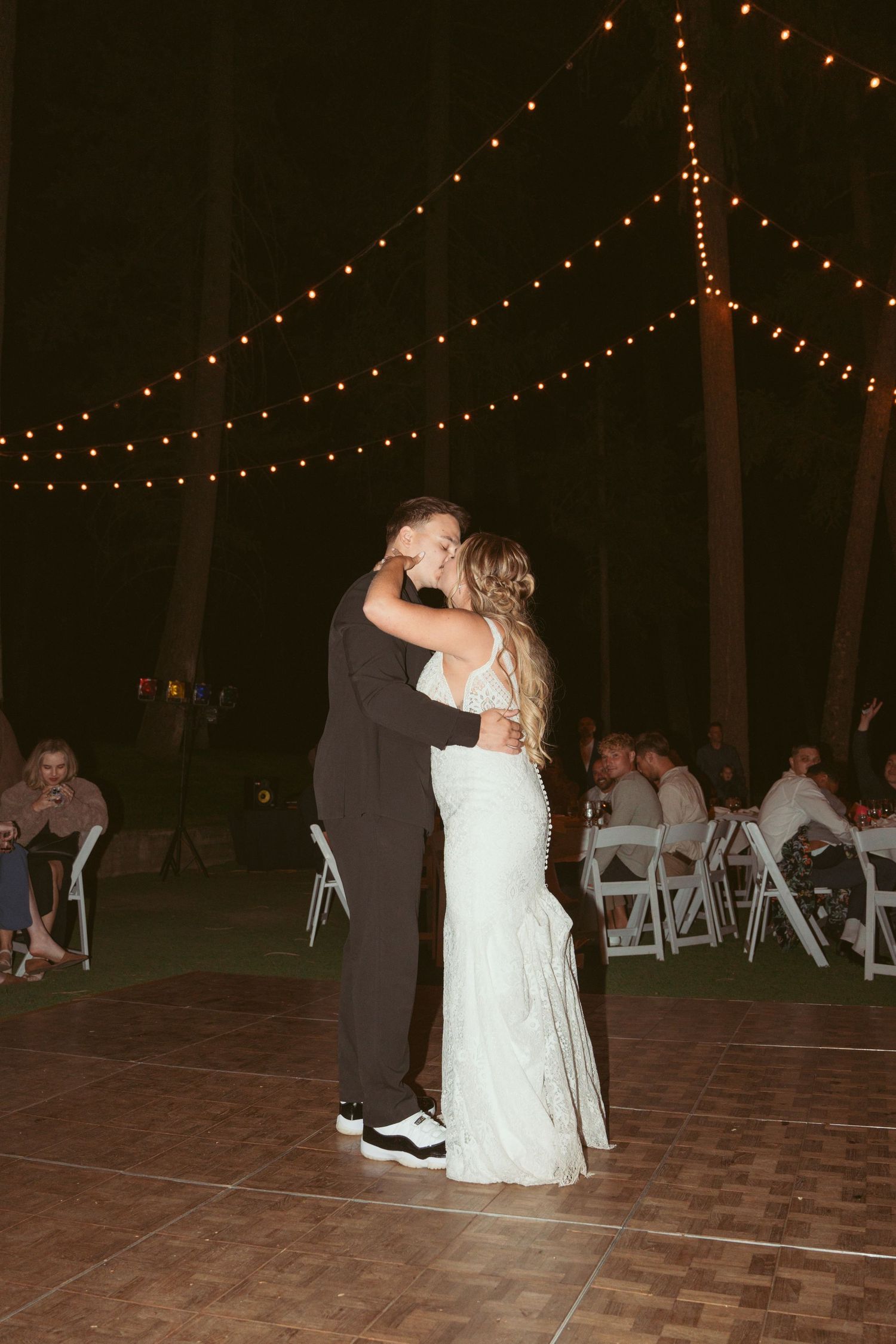 Newlyweds dancing together under twinkling lights at an evening outdoor wedding reception.