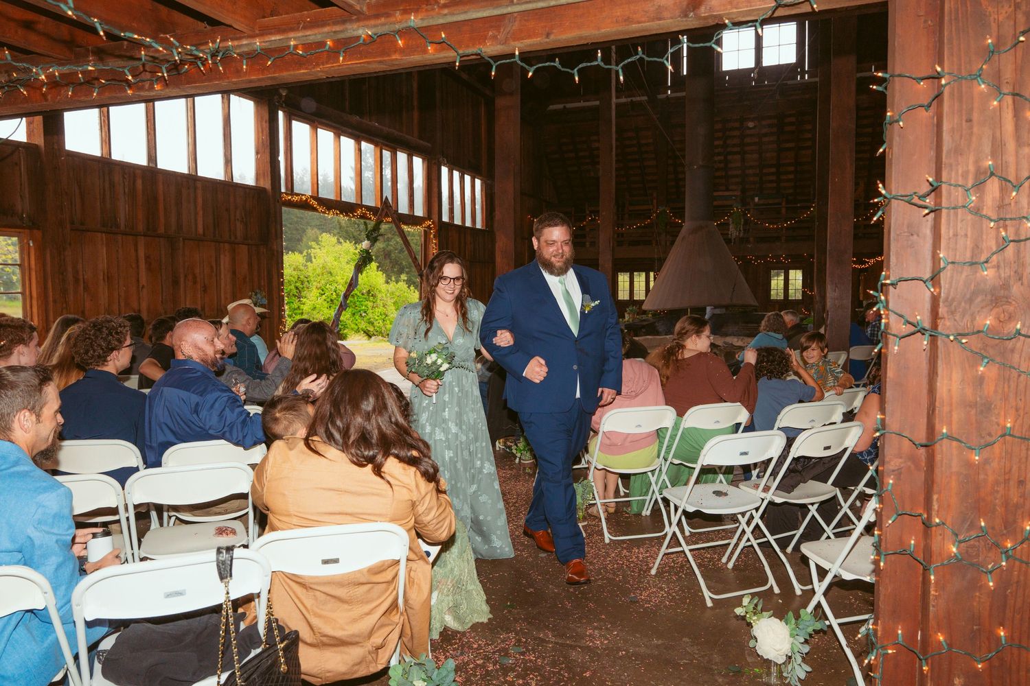 Wedding ceremony in progress inside a rustic barn with string lights and folding chair seating.