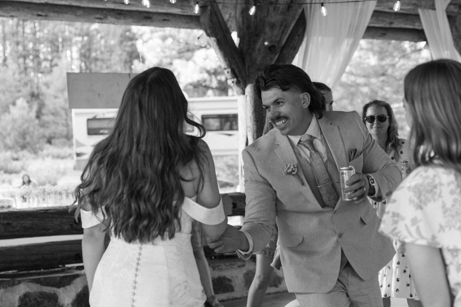 Wedding guests enjoying a candid moment under a rustic wooden pavilion in black and white.