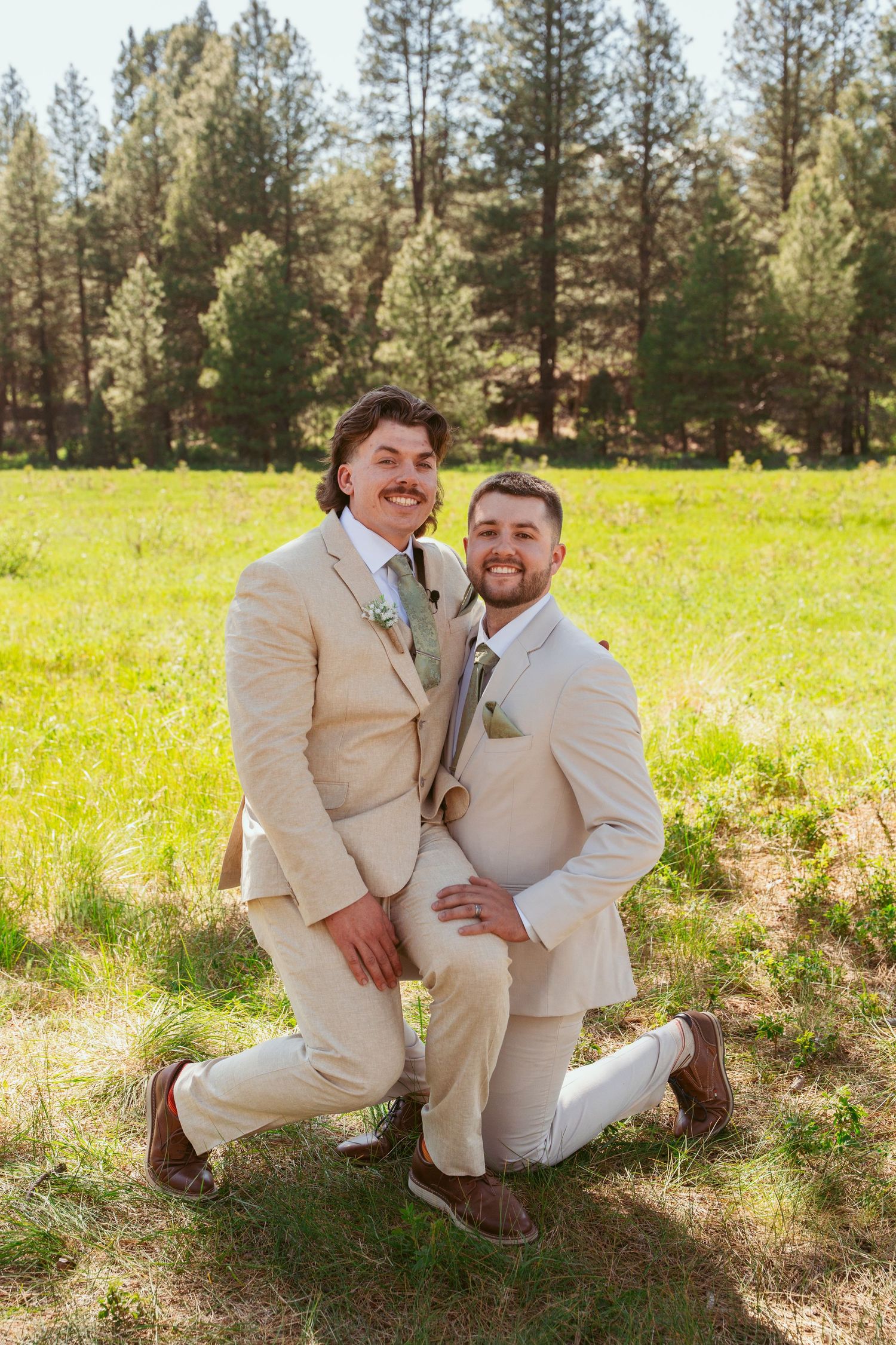 Groomsmen in cream-colored suits pose casually on grass with pine forest in background.