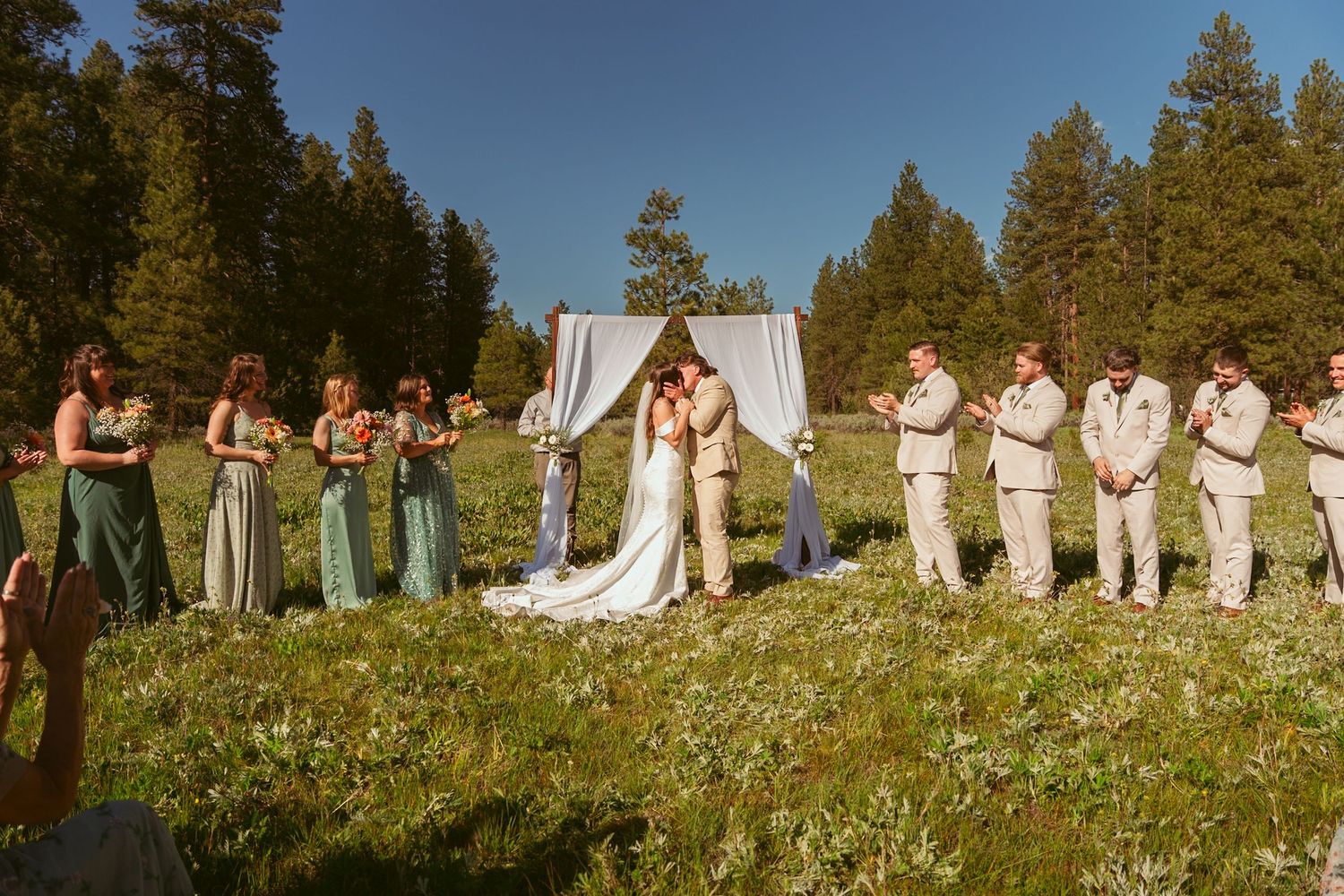 Wedding party stands in line against pine trees and white curtains in outdoor mountain ceremony setting.