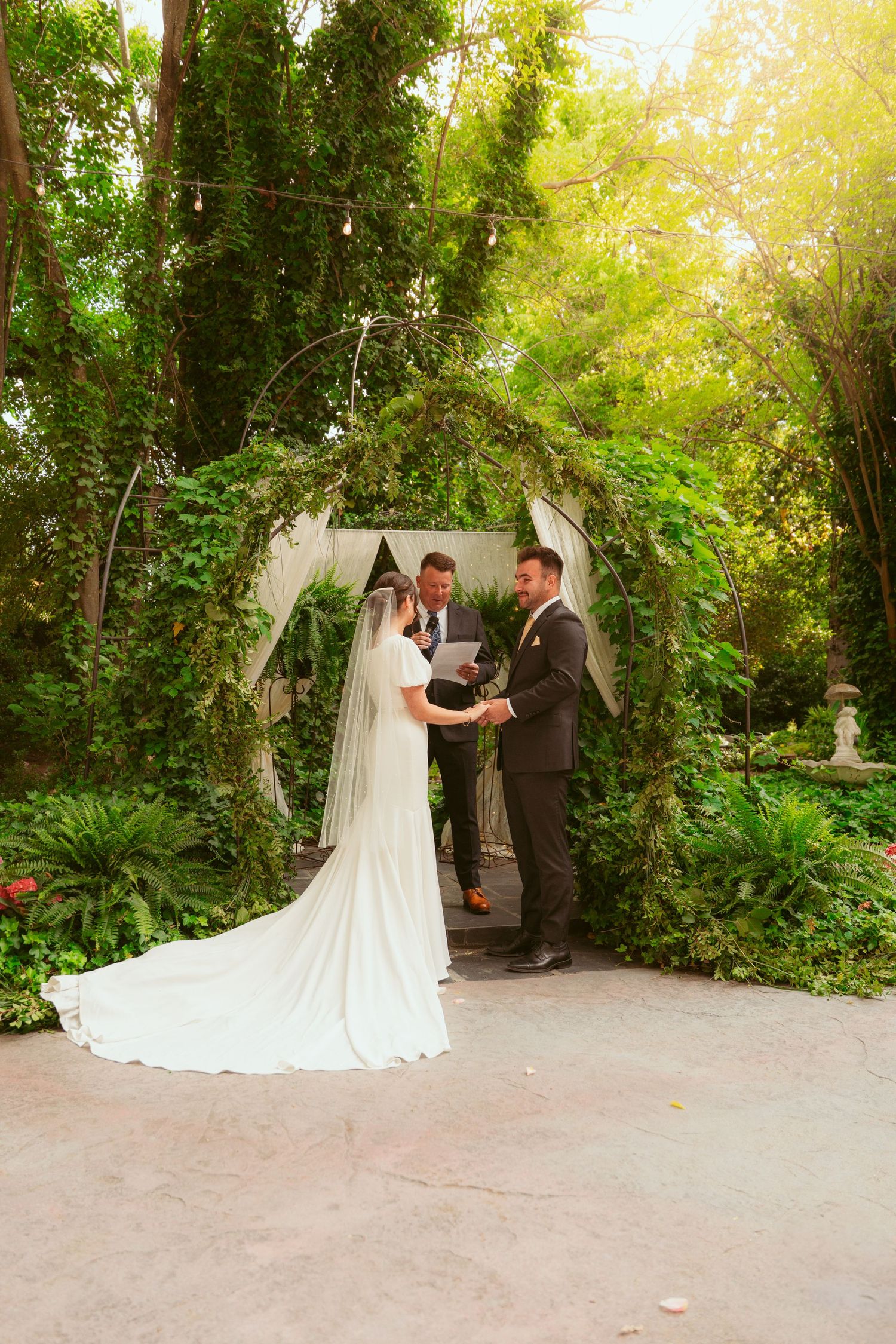 A wedding ceremony takes place under a floral arch in a sunlit garden setting.