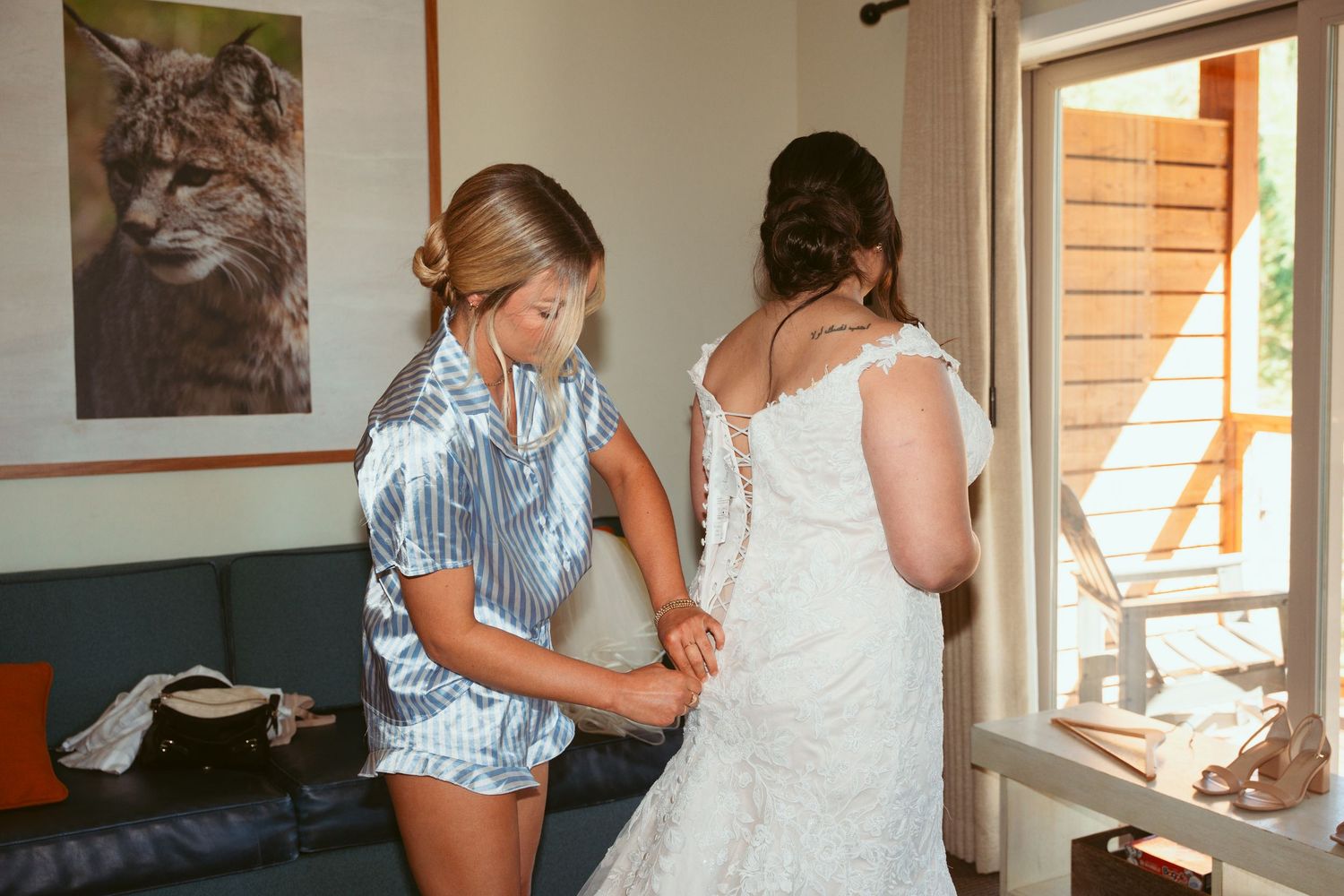 A helper assists with buttoning up a white lace wedding dress in a sunlit room with a cat portrait on the wall.