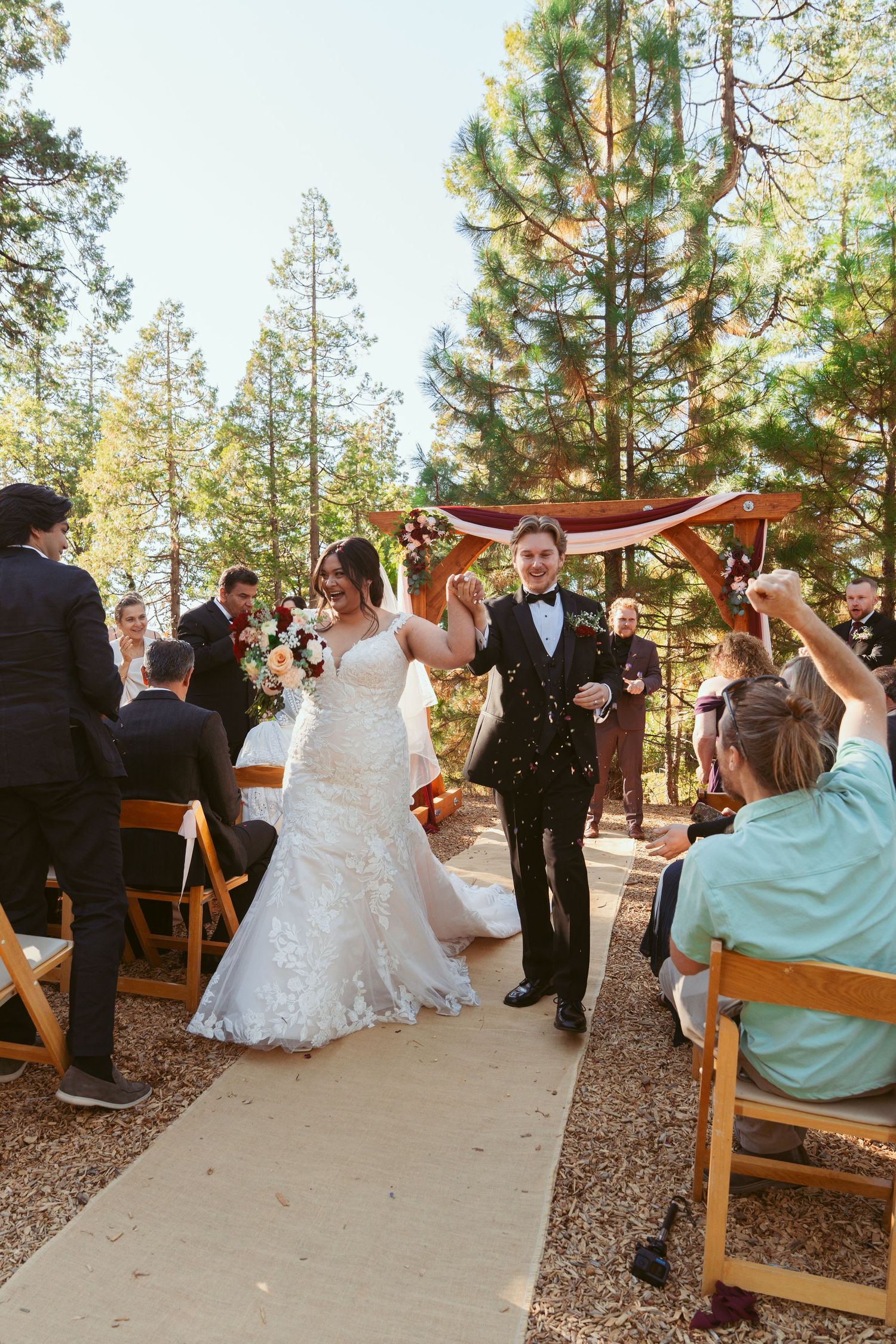 Newlyweds walk down an aisle lined with wooden chairs in a forest setting as guests celebrate their exit.