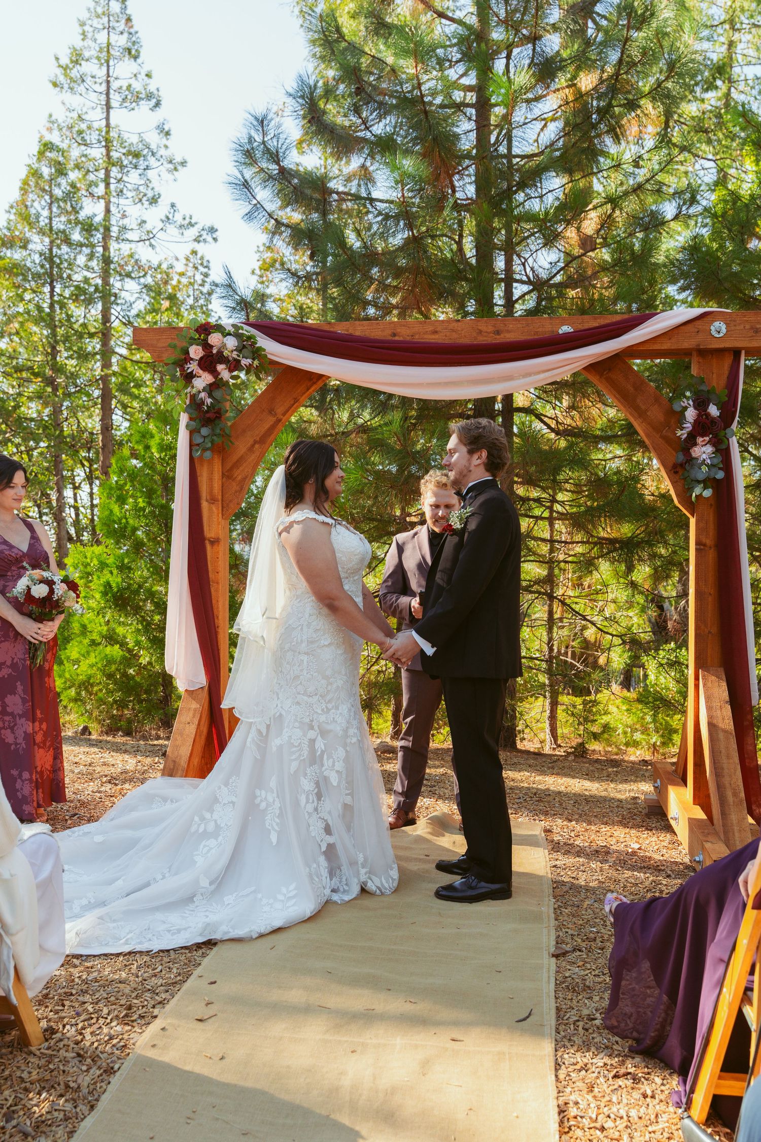 Couple exchanges wedding vows under a wooden arch decorated with burgundy fabric in a forest.