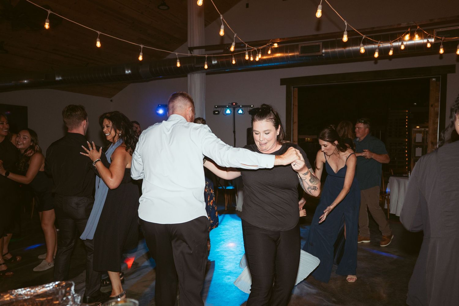 Dancers moving across a dark dance floor illuminated by overhead string lighting.
