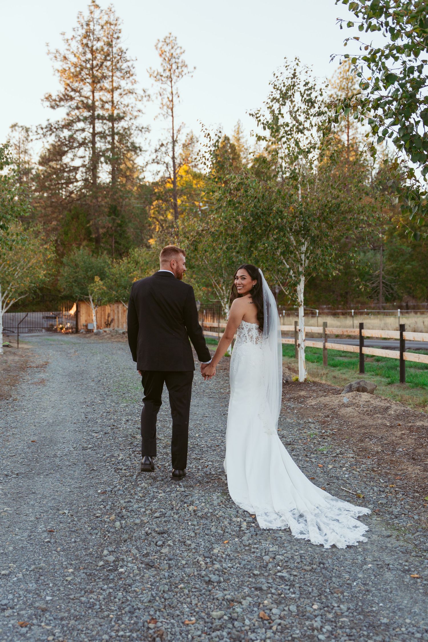 Bride in lace gown with long train walks with groom on gravel path during autumn.