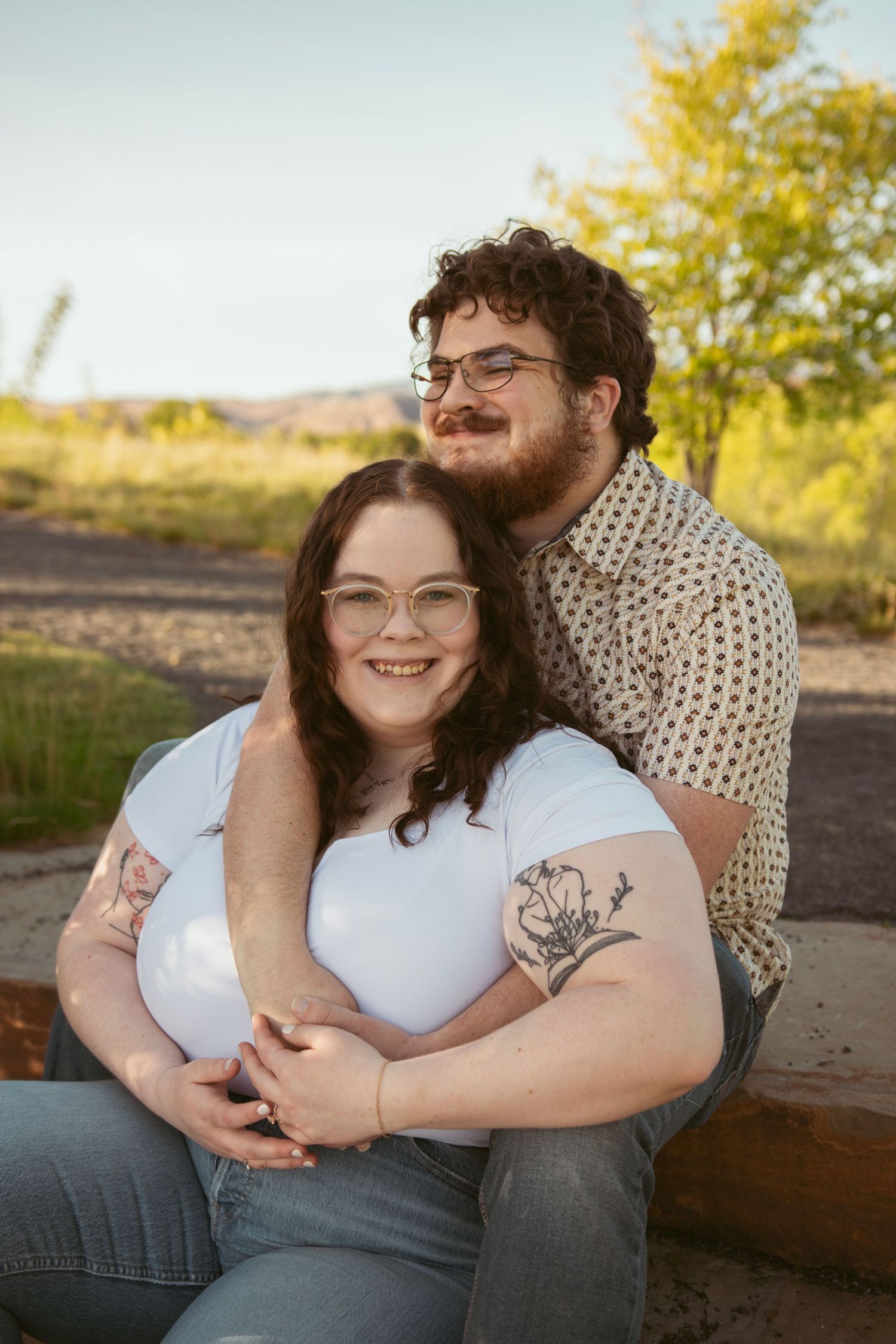 A couple embraces while sitting on wooden steps outdoors with yellow trees in background.