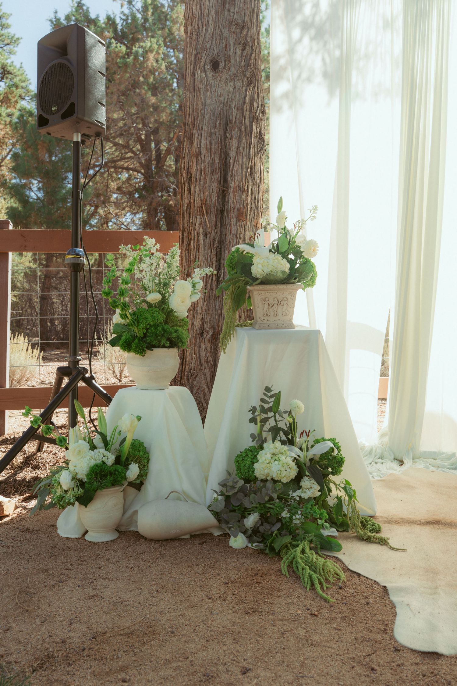 Wedding ceremony backdrop with white drapes and floral arrangements against forest trees.