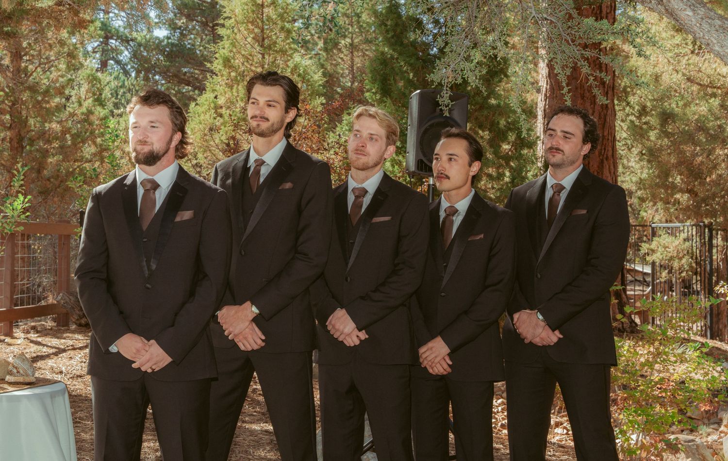 Groomsmen in dark suits pose together outdoors against forest backdrop.