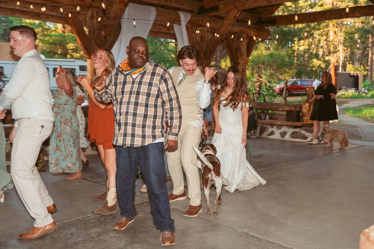 People dancing and celebrating at an outdoor wedding venue with wooden beams and string lights.