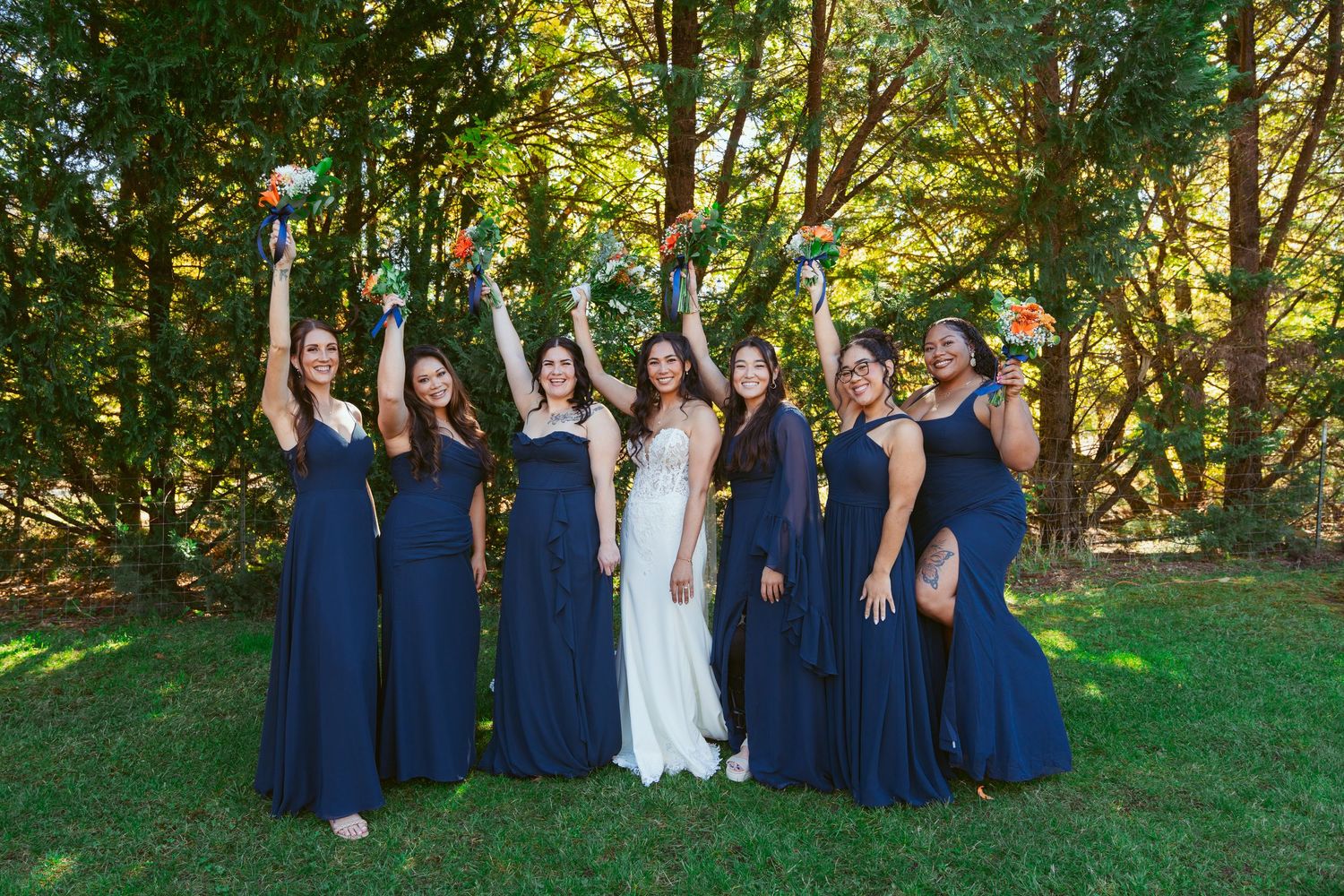 Wedding party members line up outdoors wearing navy dresses against sunlit trees.