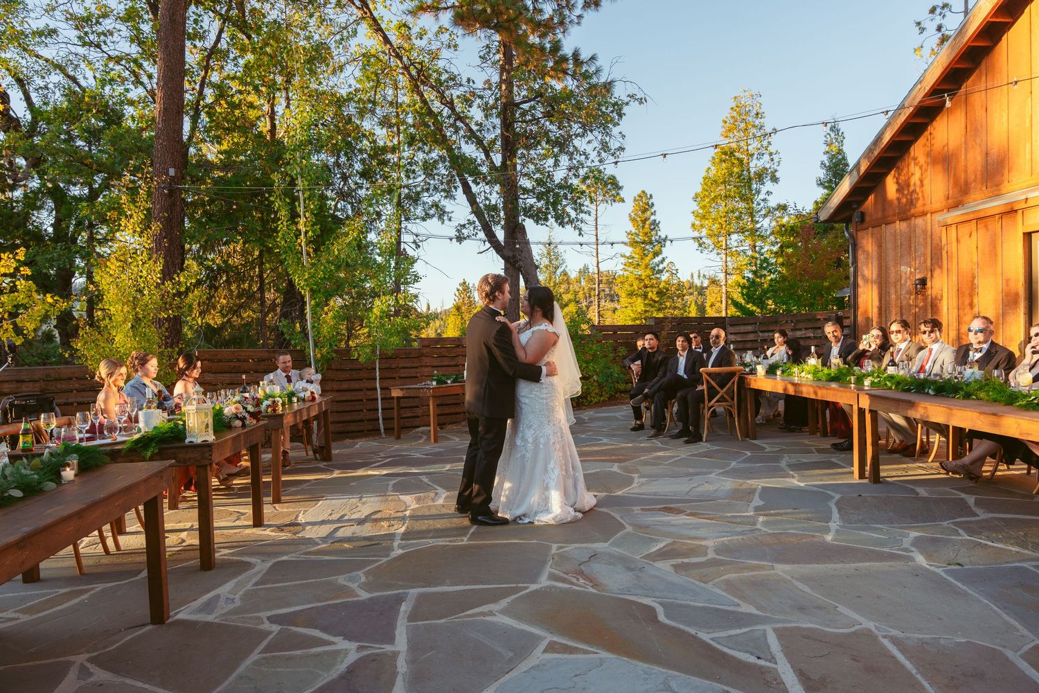 Couple dancing on an outdoor stone patio surrounded by trees during golden hour at a wedding reception.