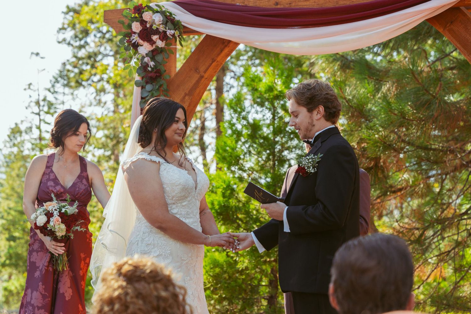Outdoor wedding ceremony features wooden arch with burgundy draping in a forest setting.