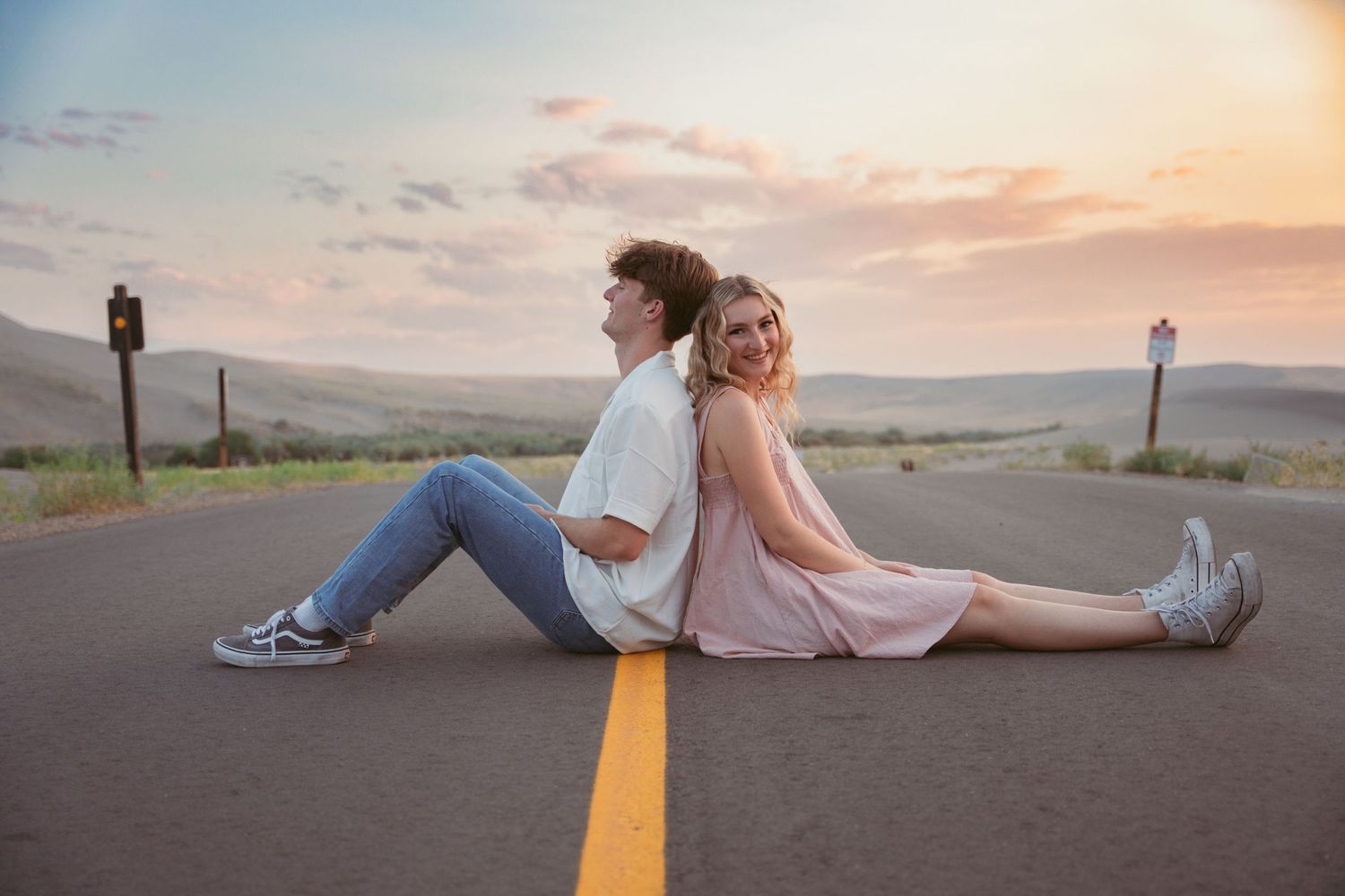 Two people sit back-to-back on a desert road at sunset.